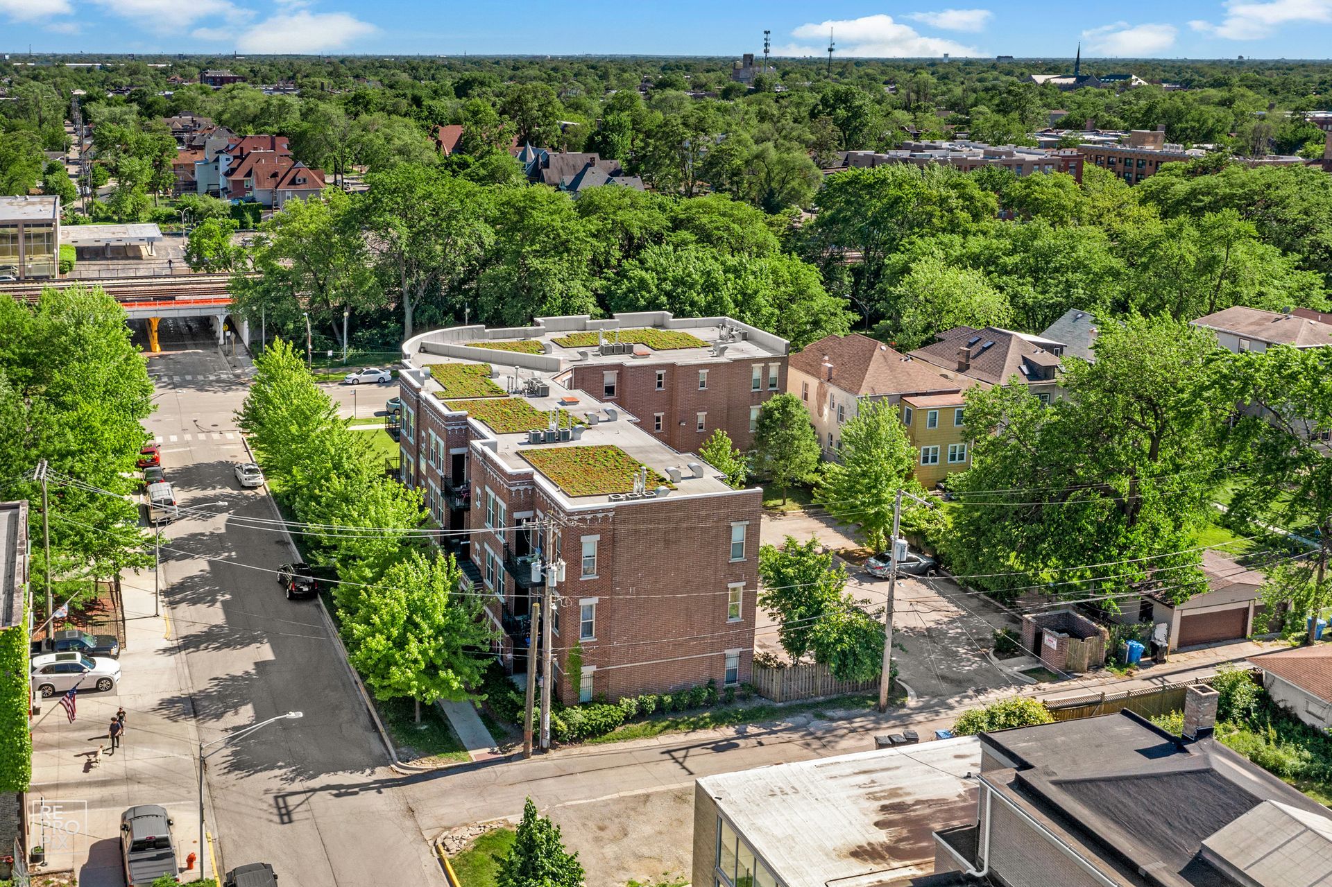 Aerial view of multi-unit brick building with green rooftop, surrounded by trees and buildings on a sunny day.