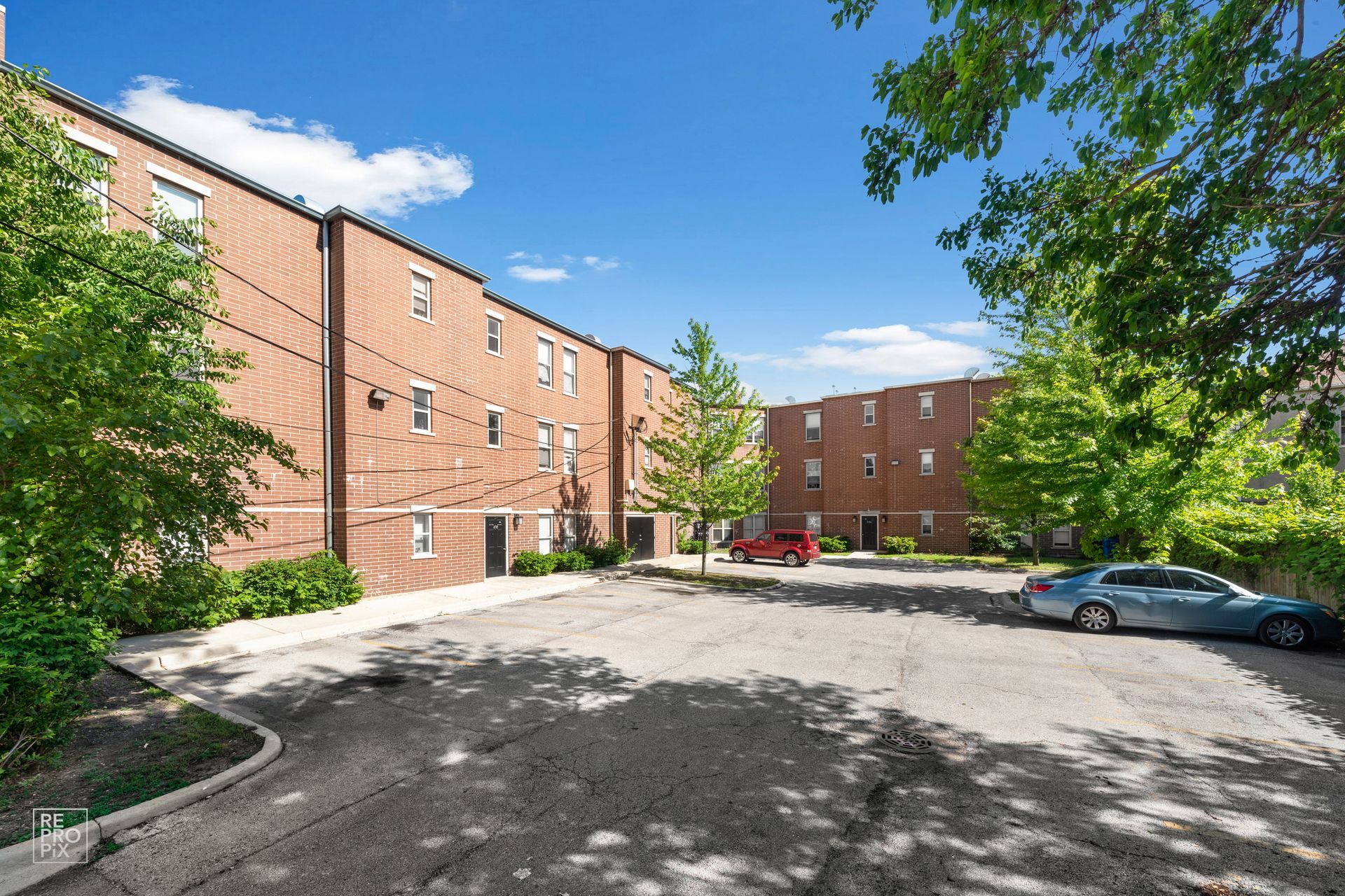 Brick apartment building with parking lot and cars on a sunny day. Trees in the foreground.