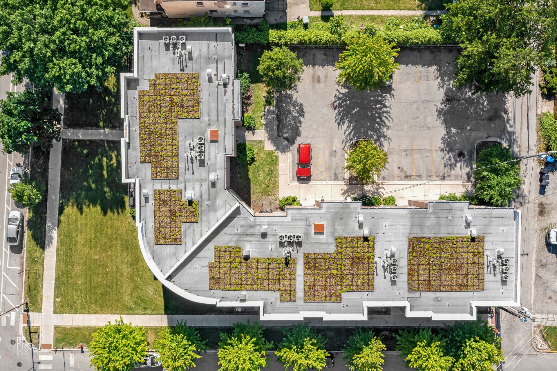 Aerial view of a green-roofed L-shaped building with a red car parked inside a parking lot surrounded by trees.