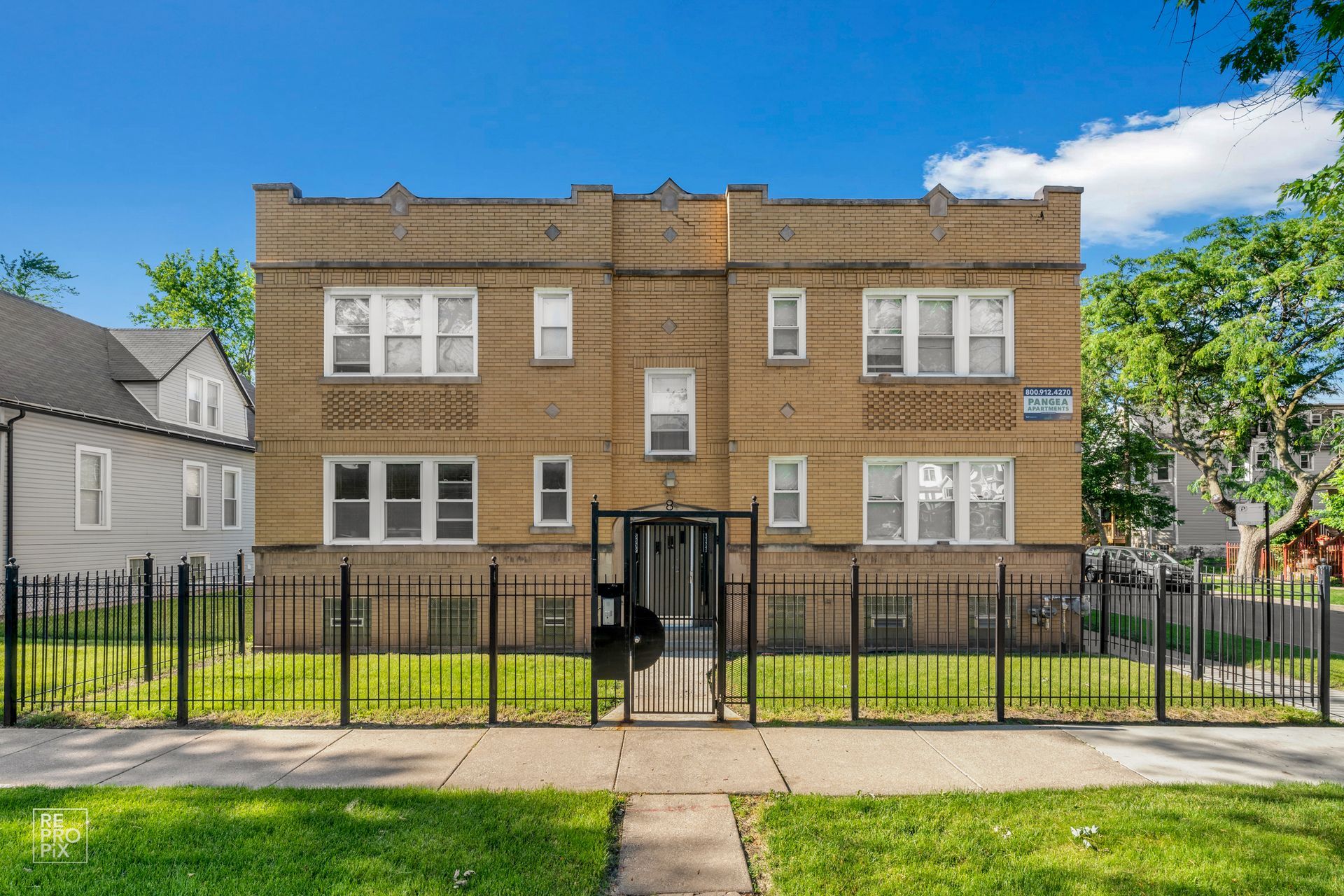 Two-story brick apartment building behind a black metal fence on a green lawn; sunny day.