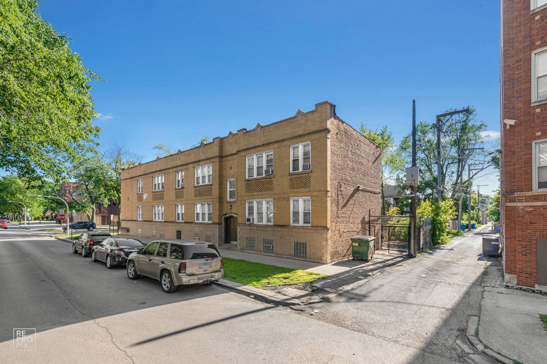 Multi-unit brick apartment building on a residential street; parked cars and a green trash bin visible.