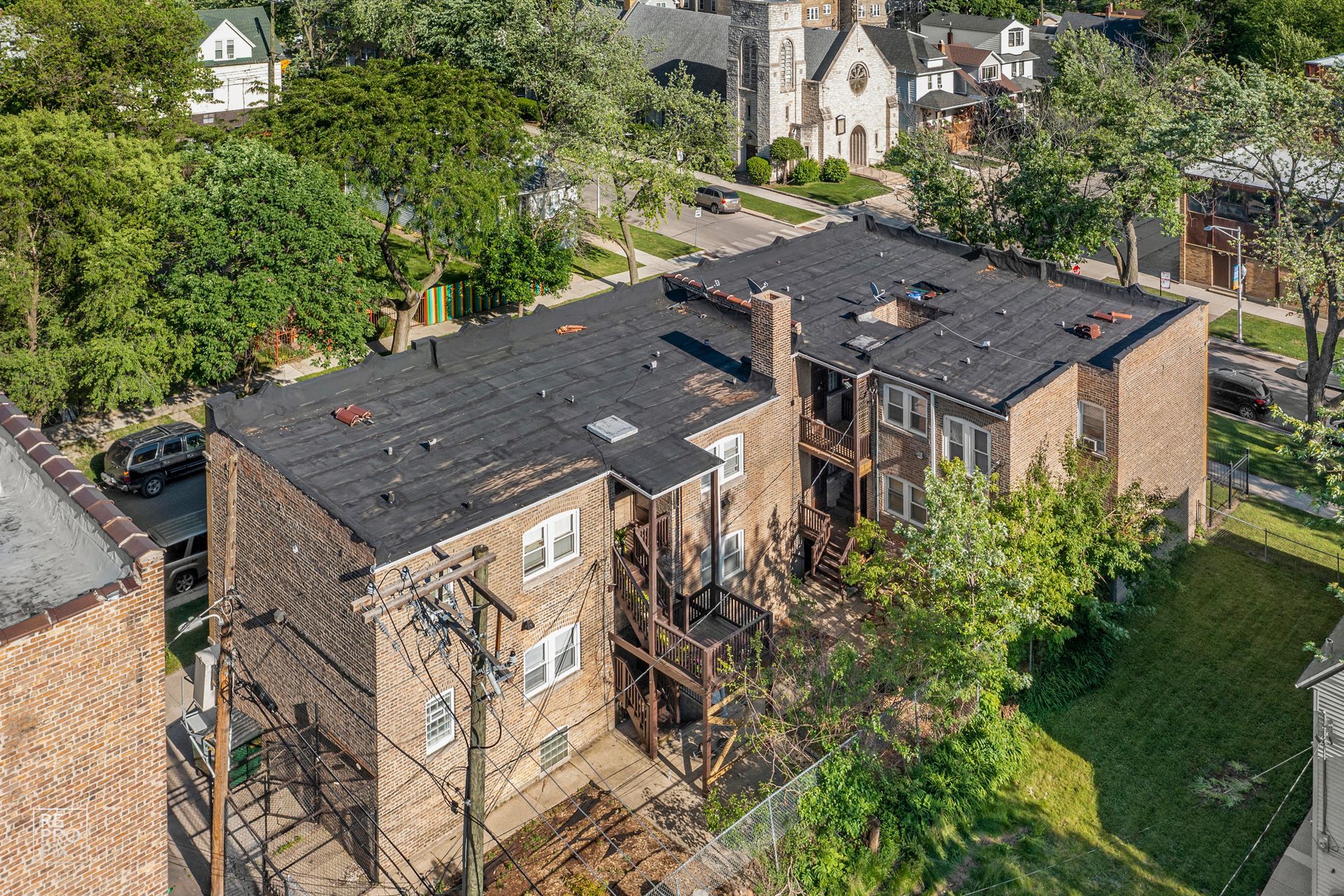 Aerial view of a brick apartment building with a dark roof surrounded by trees and a church.