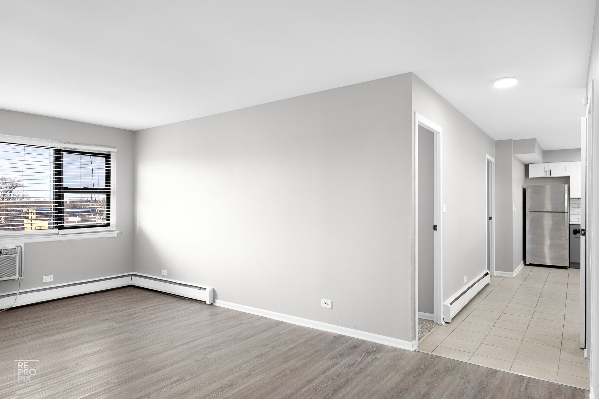 Empty apartment interior with light grey walls, hardwood floors, and a kitchen visible in the hallway.