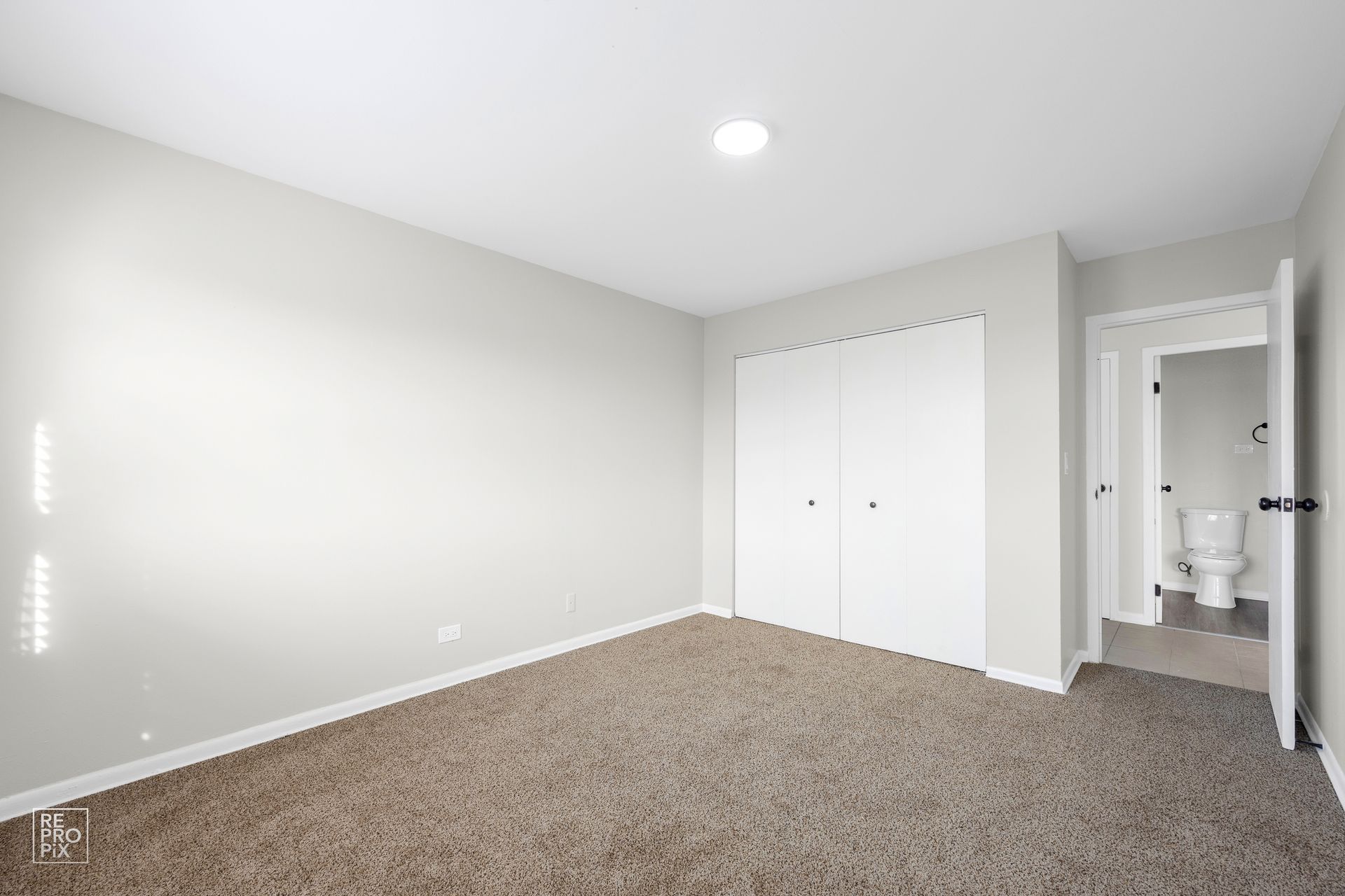 Empty bedroom with light brown carpet, white closet doors, and a doorway to a bathroom.