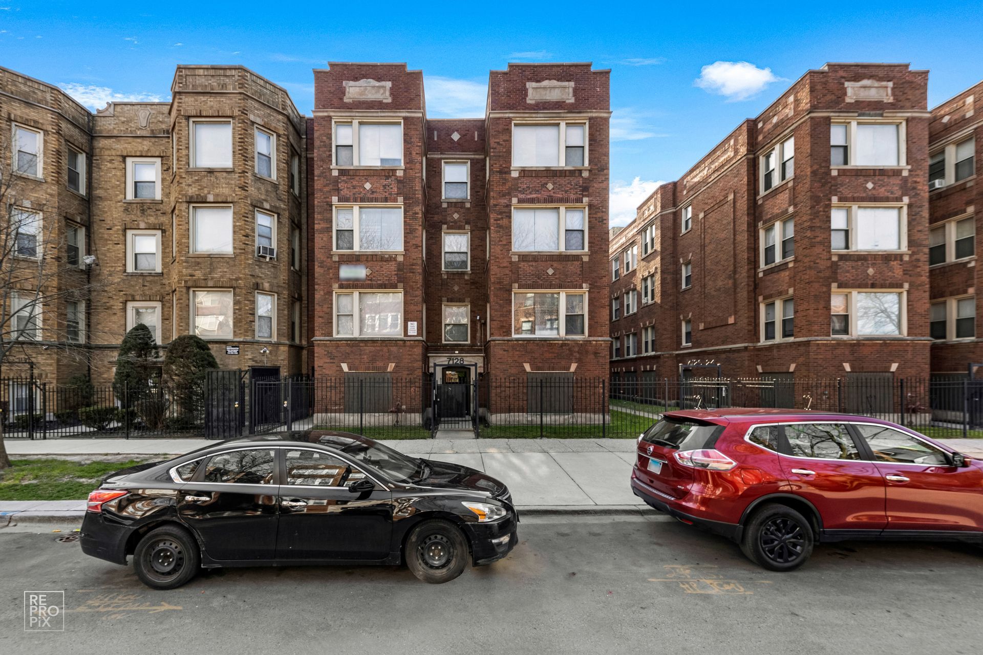 Row of brick apartment buildings with cars parked in front on a city street under a partly cloudy sky.
