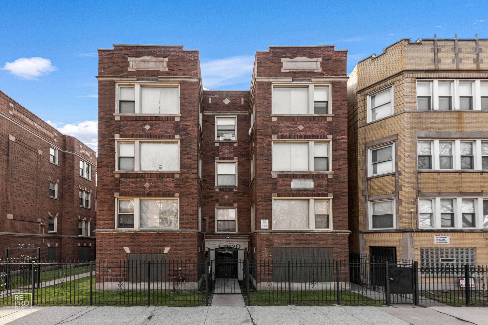 Brick apartment building with windows, a dark metal fence, and a blue sky.