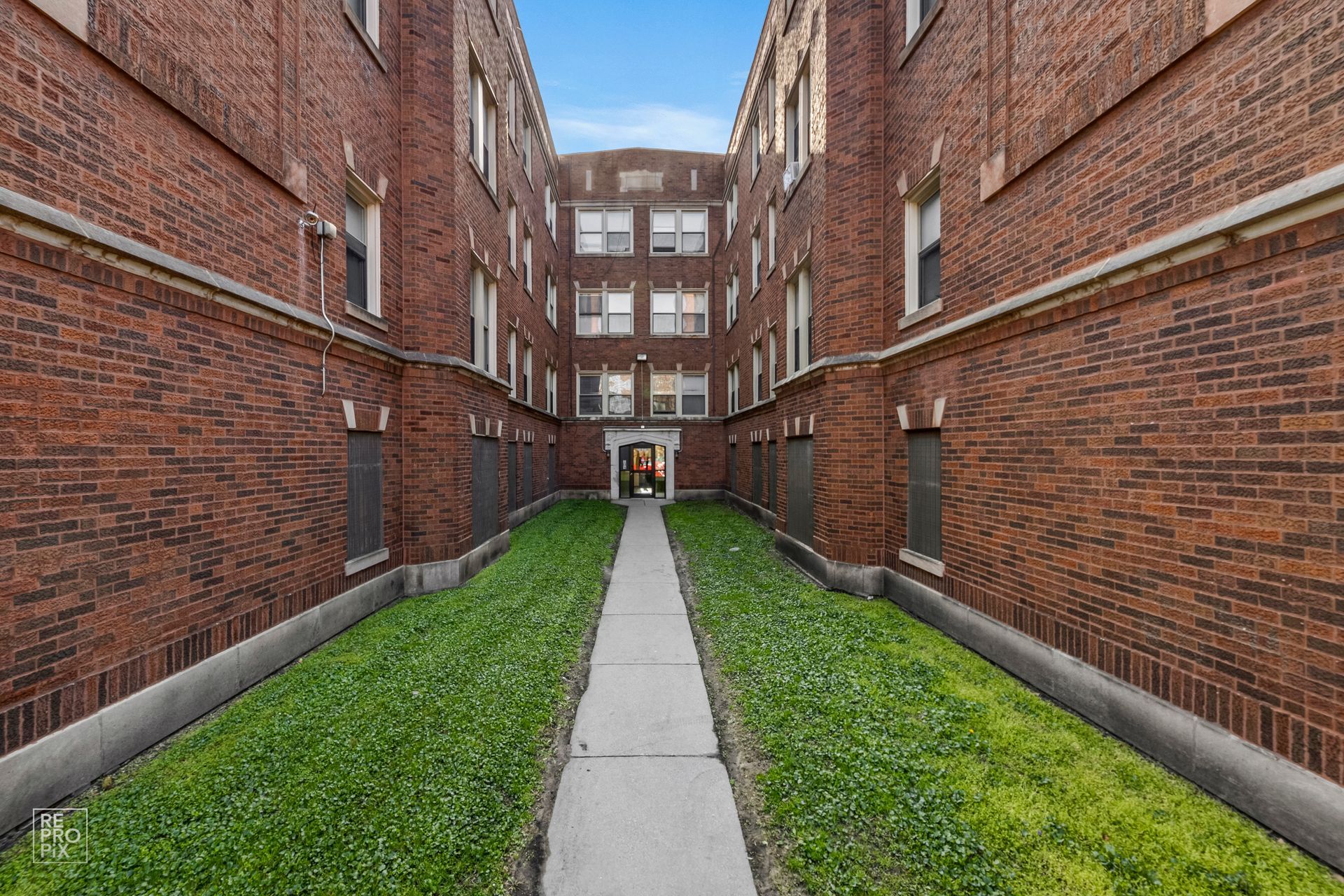 Row of brick apartment buildings with a sidewalk, street, cars, and black metal fences under a blue sky.