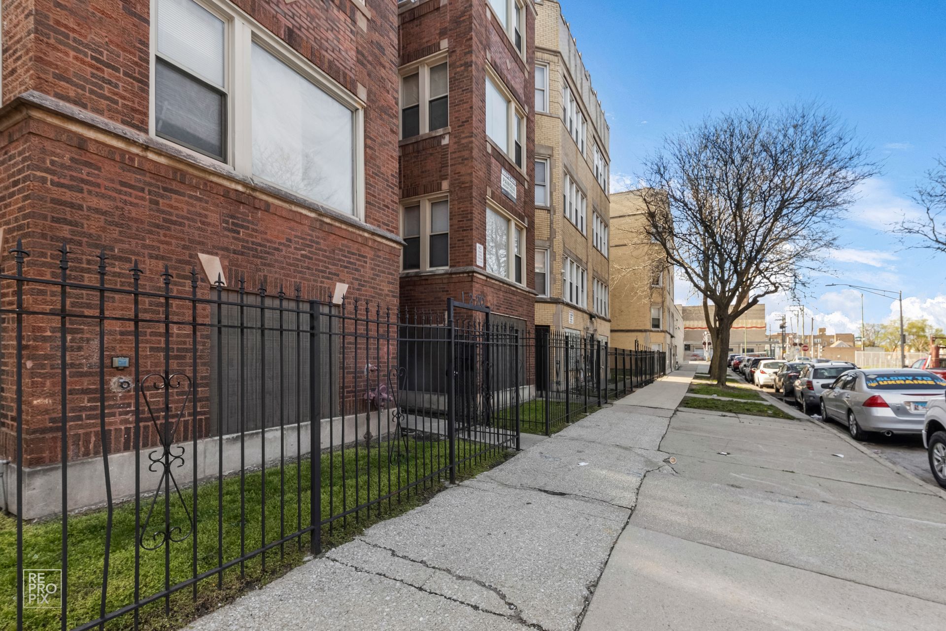 Brick apartment buildings with a sidewalk, metal fence, and parked cars on a sunny day.