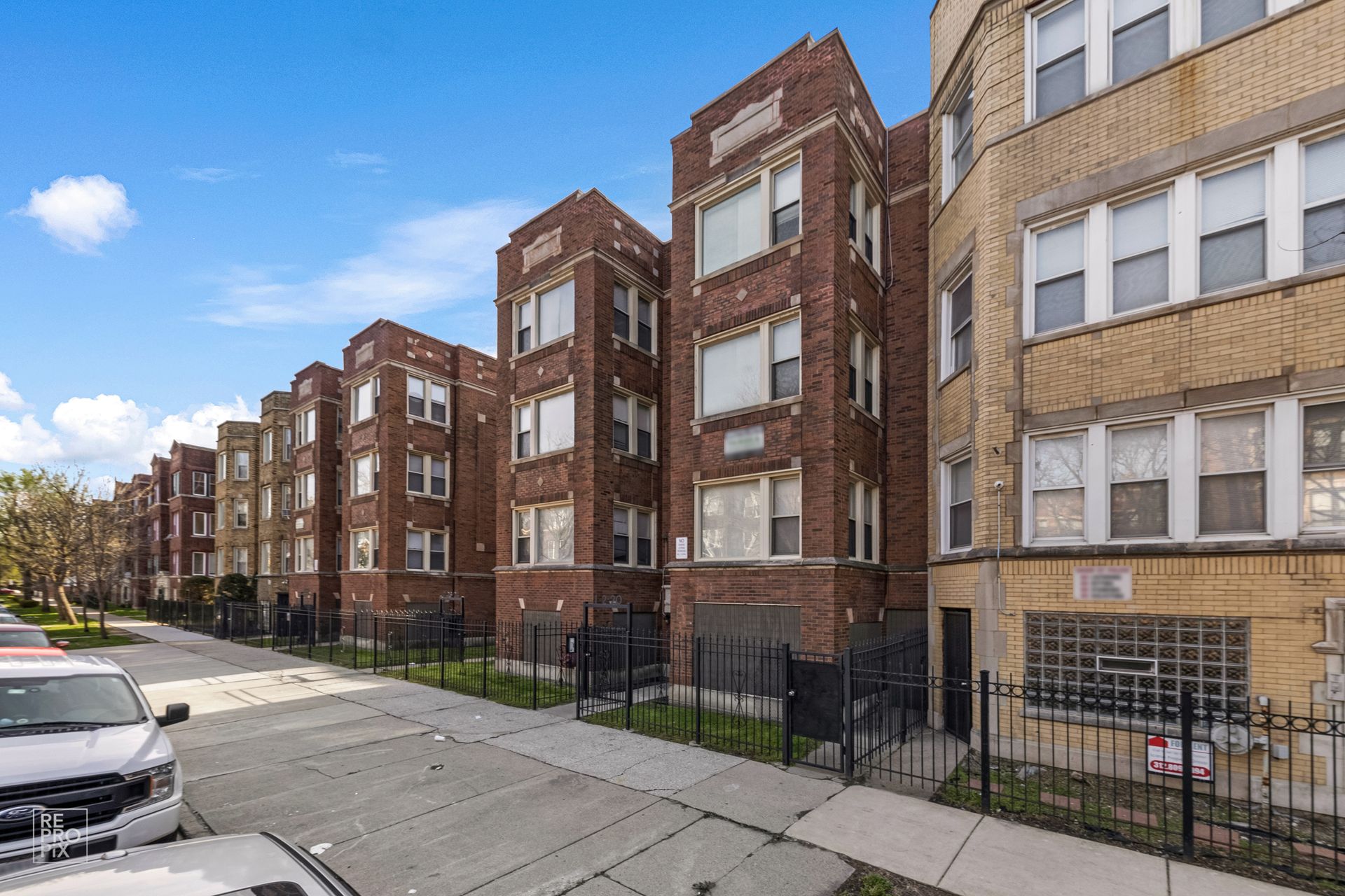 Row of brick apartment buildings with dark fences and parked cars on a sunny day.