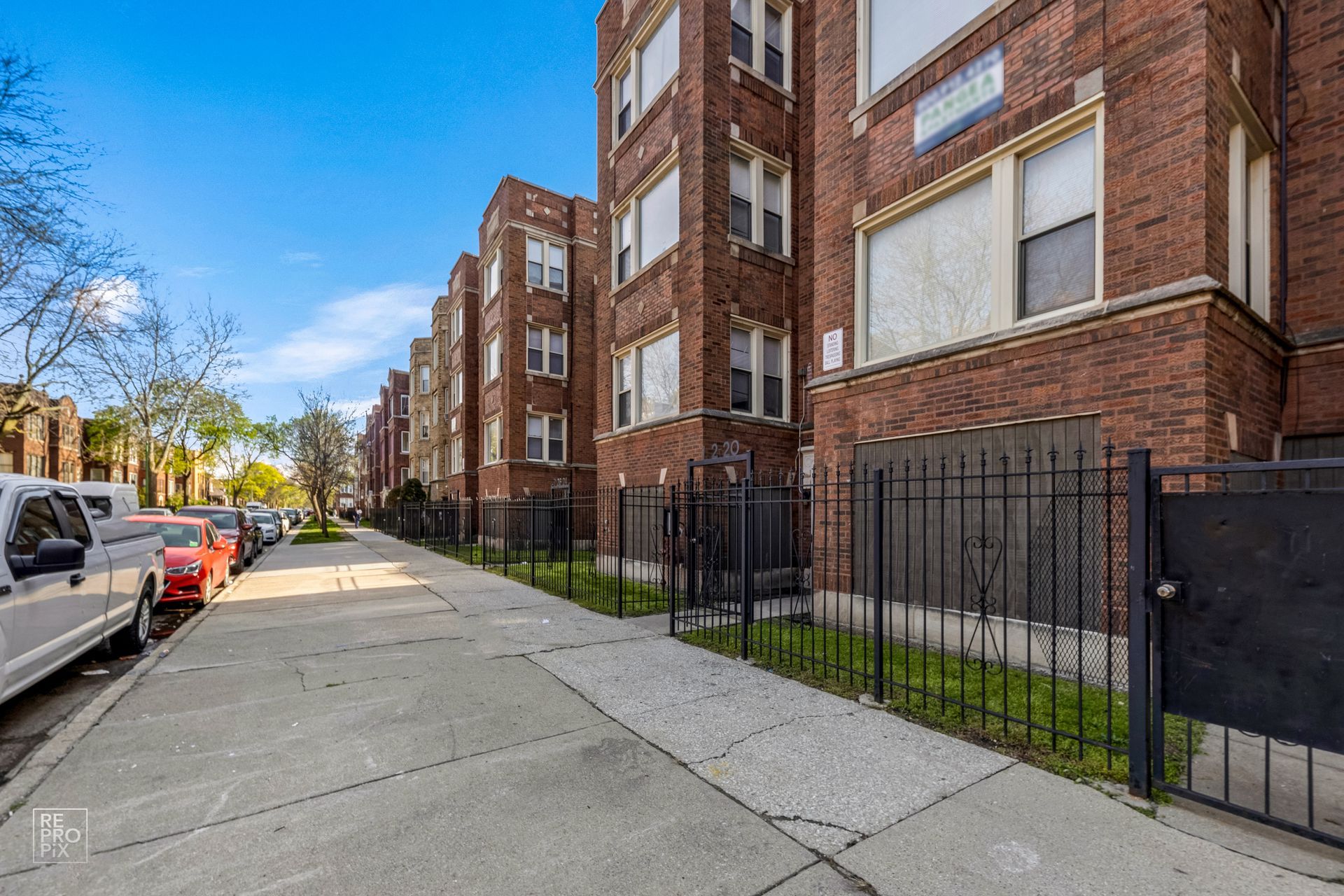 Row of brick apartment buildings along a sidewalk with parked cars and a metal fence.