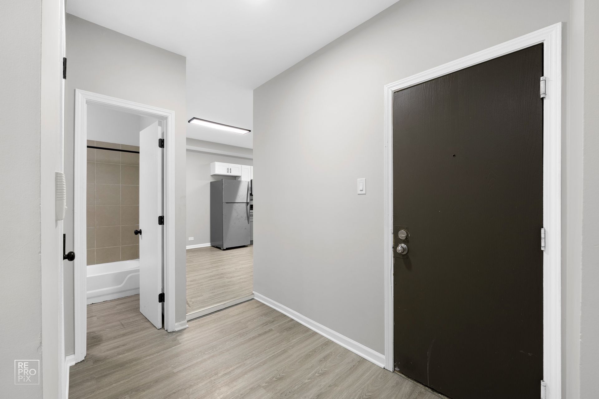 Hallway with light gray walls and wood-look flooring. Opens to bathroom on left and kitchen ahead. Dark brown door on right.