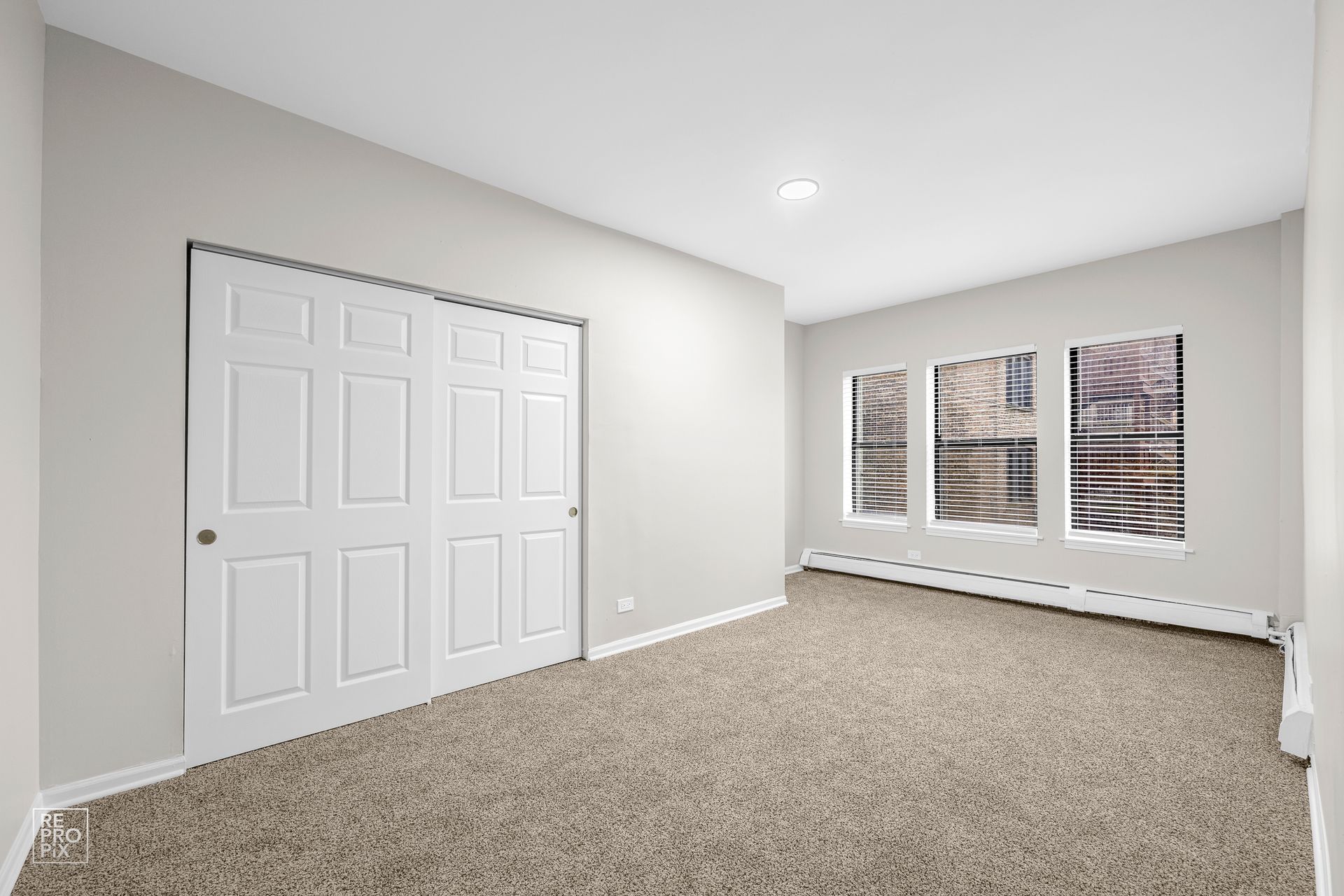 Empty room with white walls, beige carpet, closet doors, and windows with a city view.