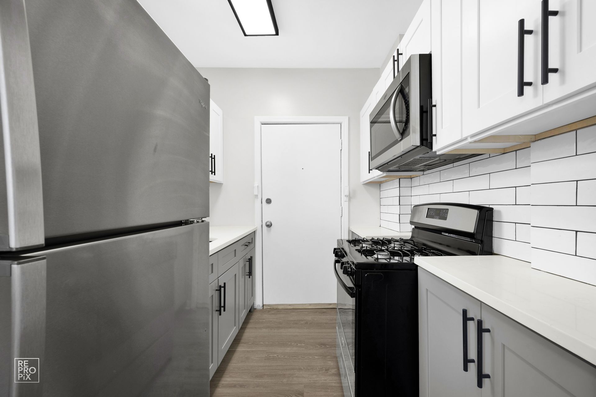 Narrow kitchen with stainless steel fridge, grey and white cabinets, black appliances, and a door at the end.