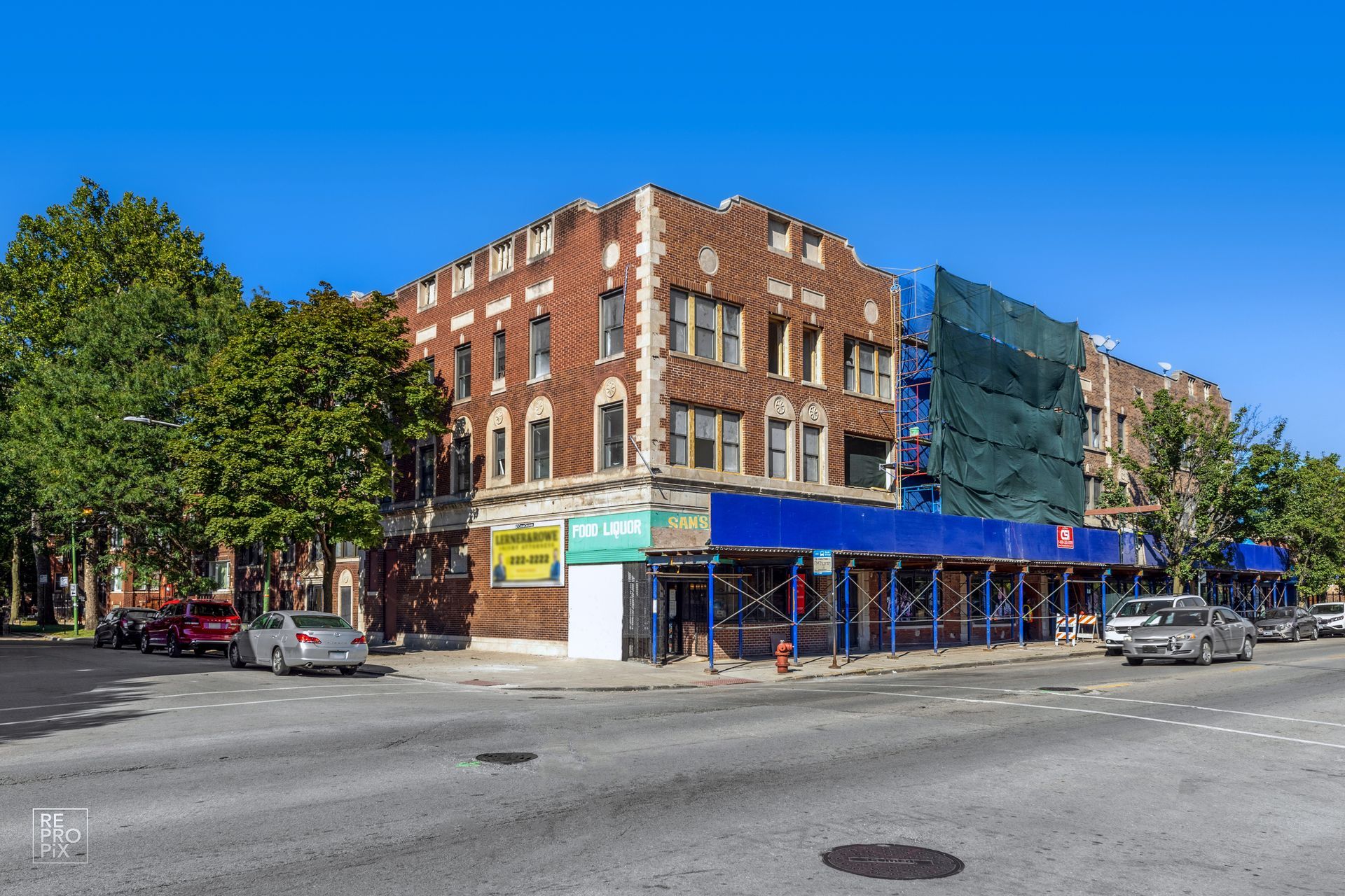 Brick building on a corner with scaffolding and a blue awning. Cars parked on the street.