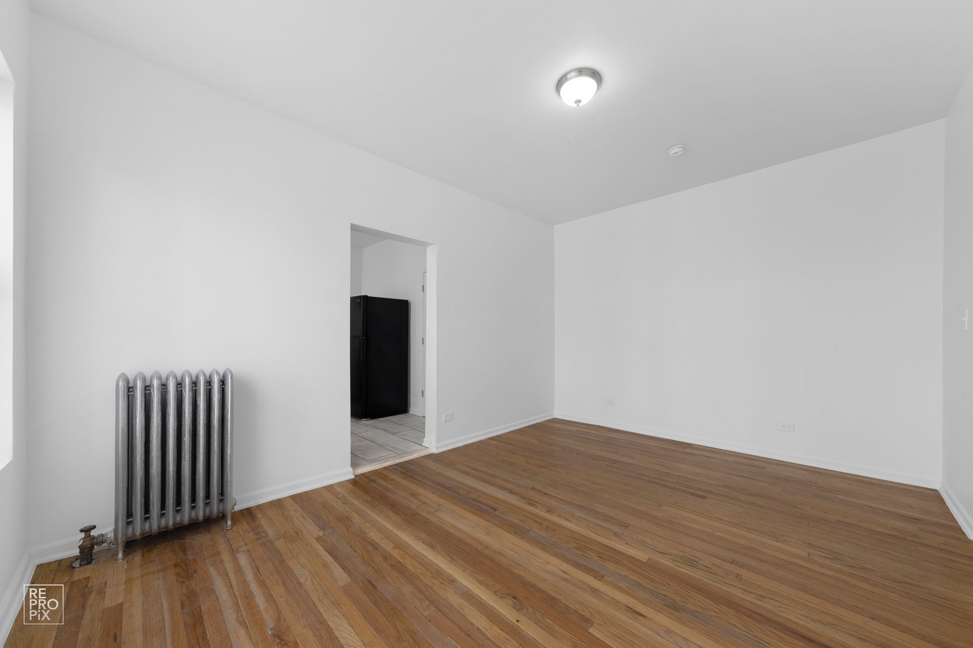 Empty room with hardwood floors, white walls, radiator, and doorway to another room.