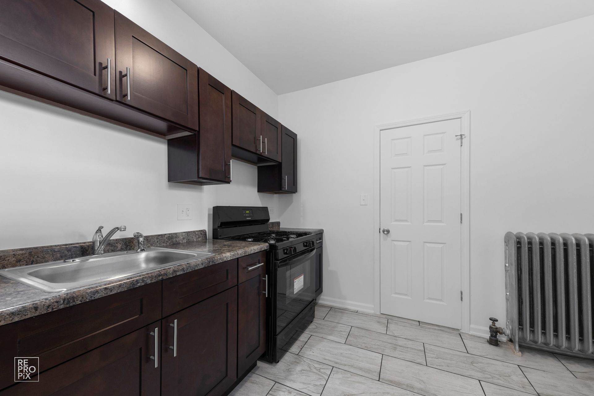 Dark wood kitchen cabinets, stainless steel sink, black stove, white walls, white door, and gray tile floor.