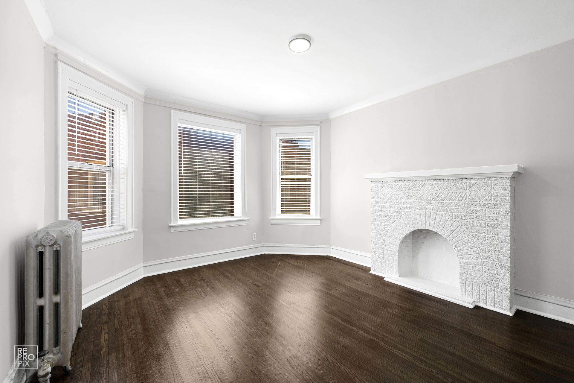 Empty, bright room with bay windows, dark wooden floor, fireplace, and radiator.