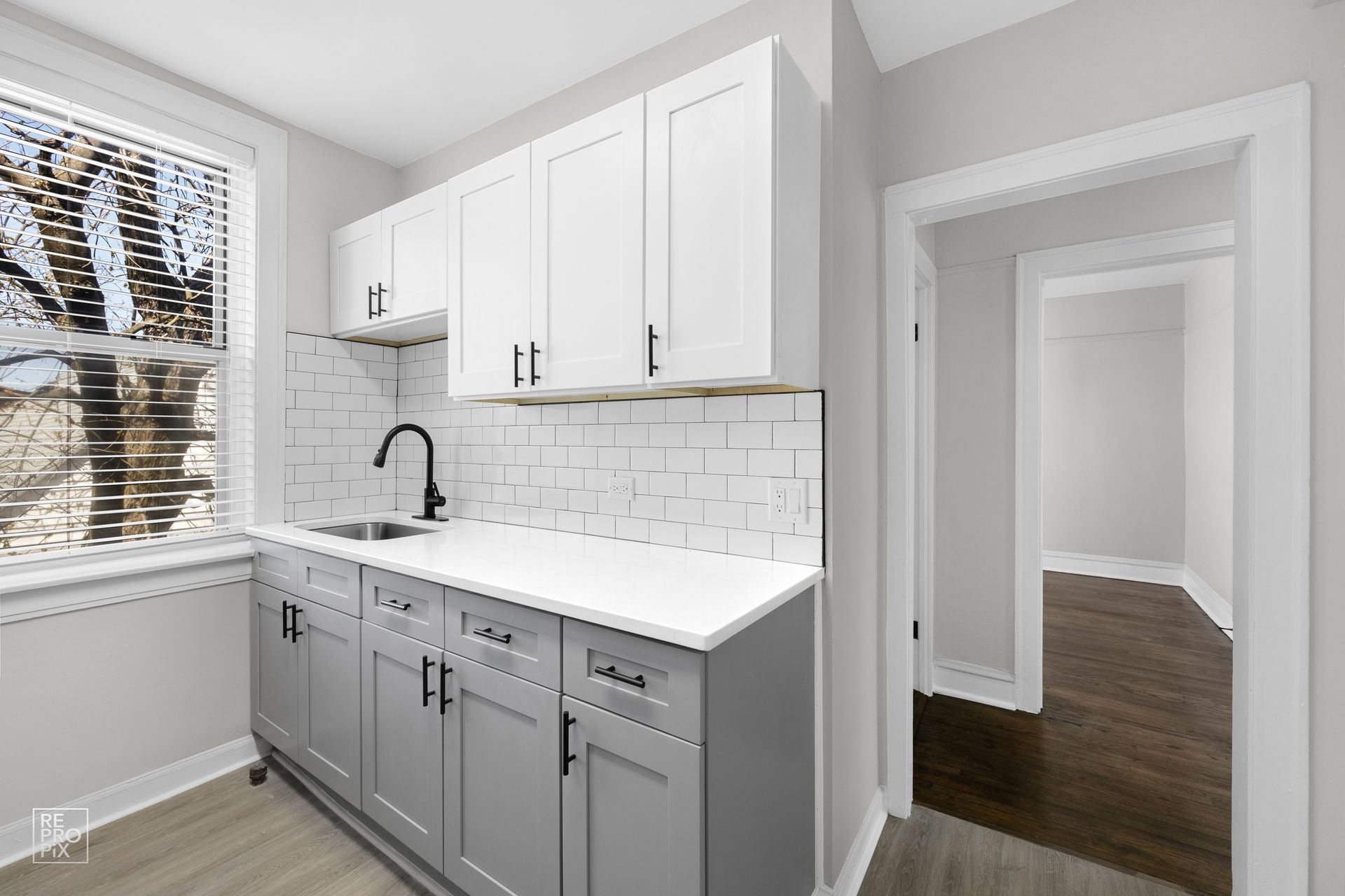 Kitchenette with white and gray cabinets, sink, and window. Doorway leads to a room.
