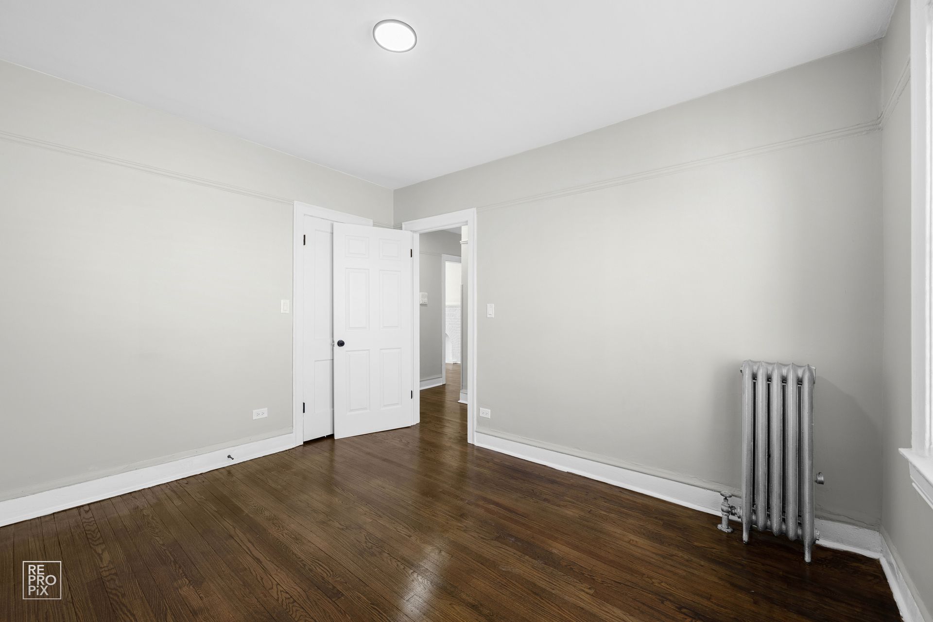 Empty room with hardwood floor, closet doors, radiator, and a doorway to another room.