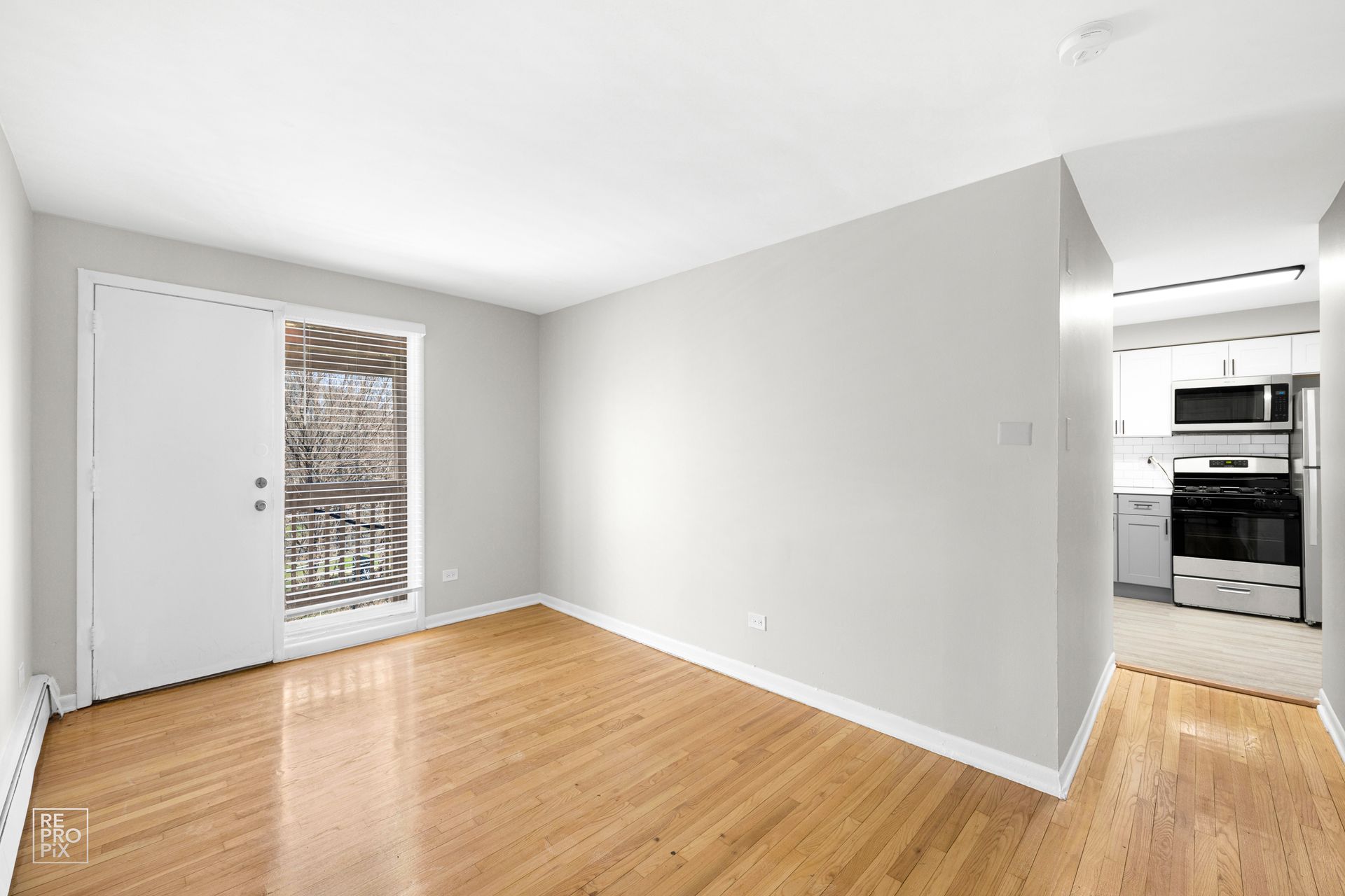 Empty apartment interior with wood flooring, light gray walls, and a doorway to a kitchen.