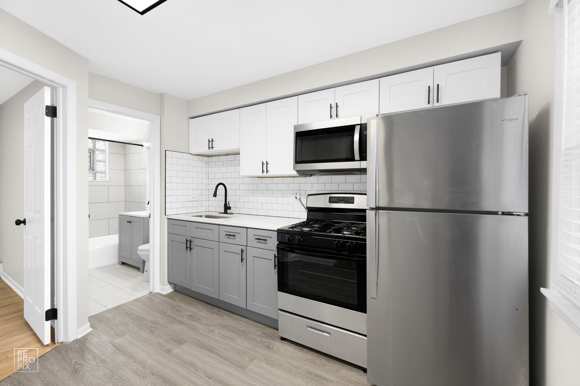 Modern kitchen with gray and white cabinets, stainless steel appliances, and a glimpse of a bathroom.
