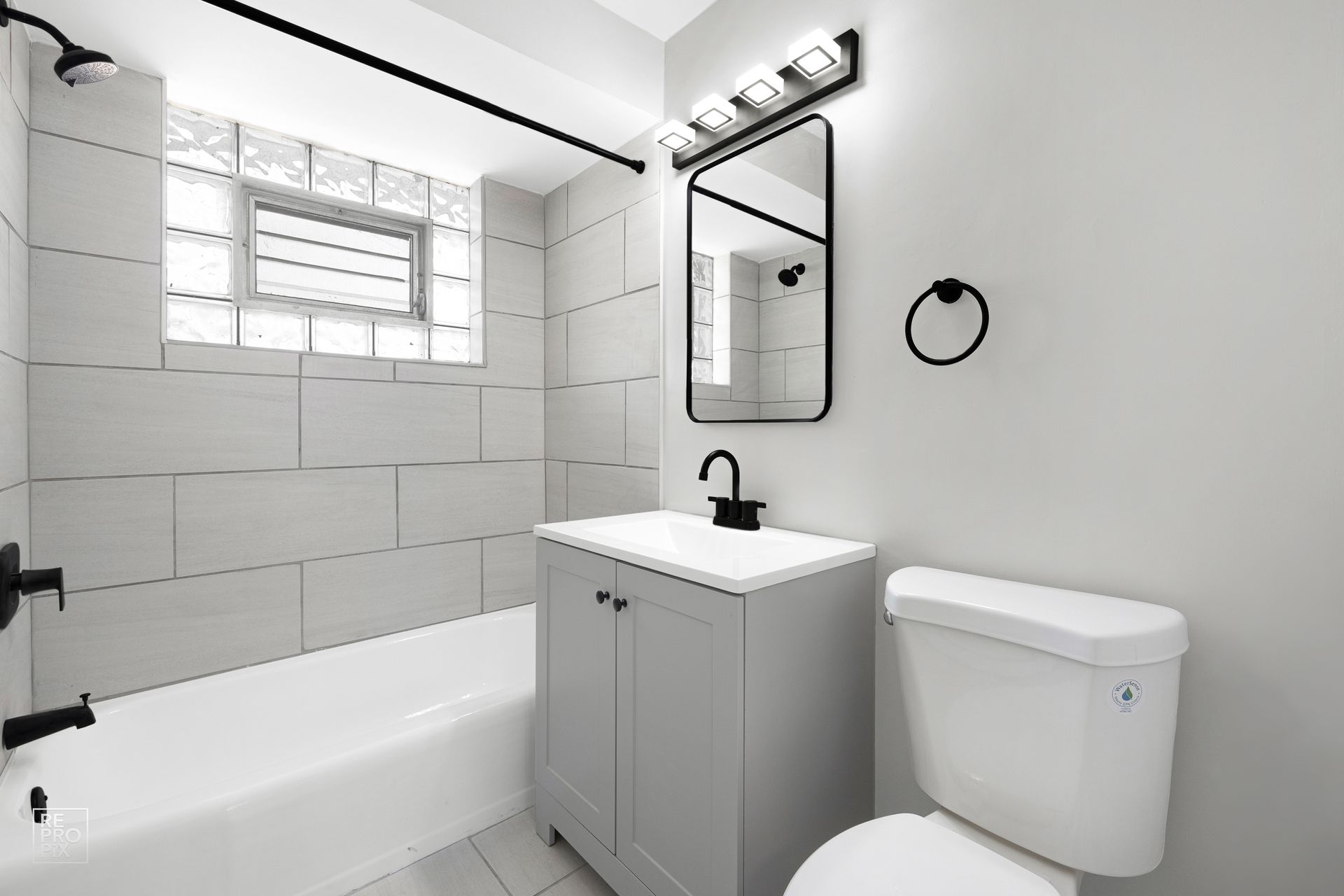 Modern bathroom with white and gray tiles, gray vanity, black fixtures, and a rectangular mirror.