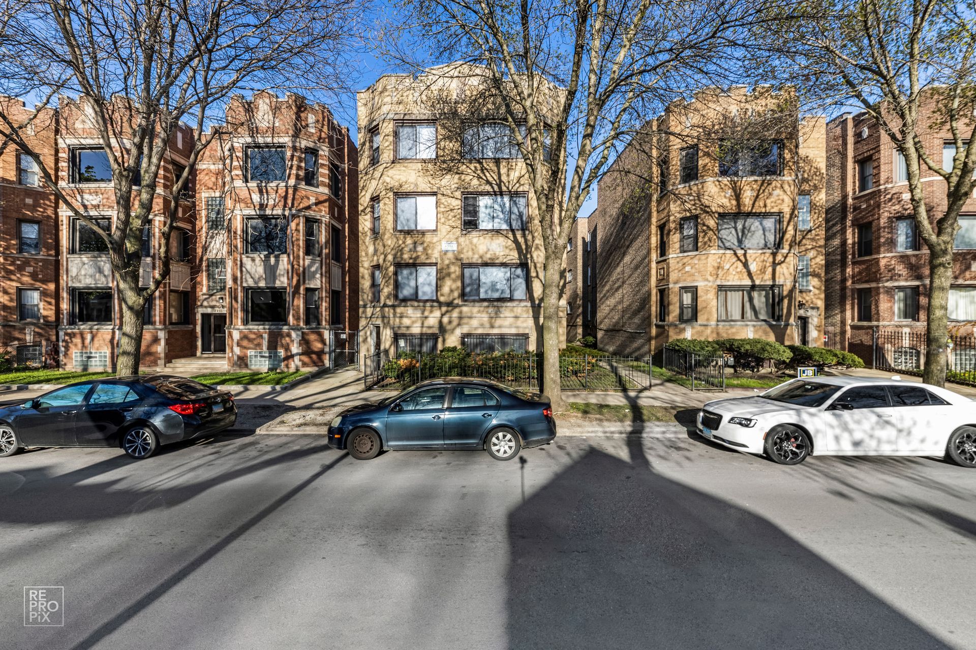 Three brick apartment buildings with cars parked on the street on a sunny day.