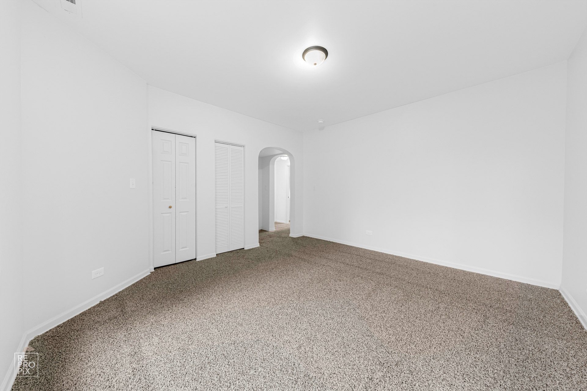 Empty room with white walls, carpeted floor, and recessed lighting. Features two doors and an arched doorway.
