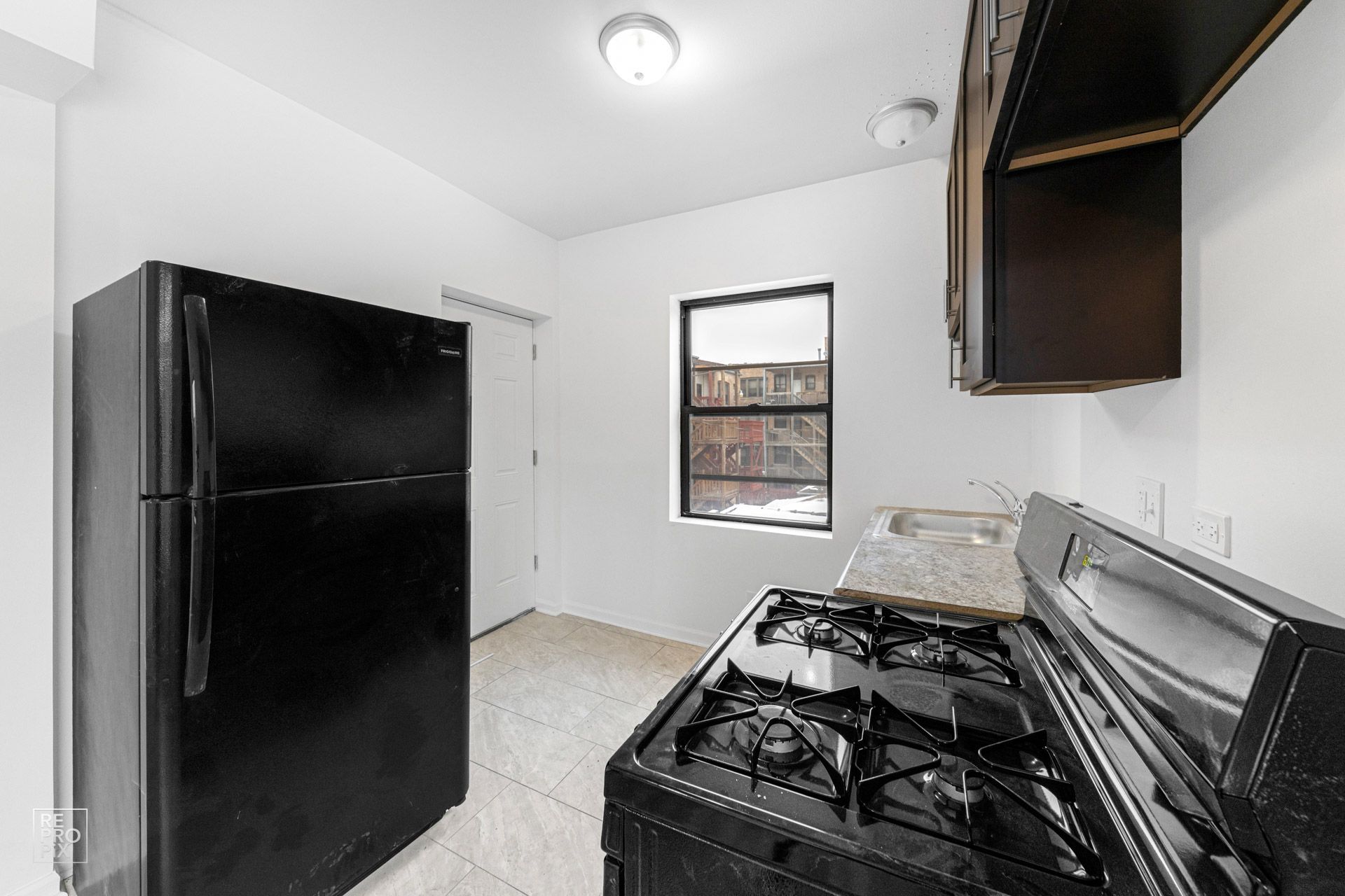 Black refrigerator, gas stove, kitchen window, and white walls in a small kitchen.