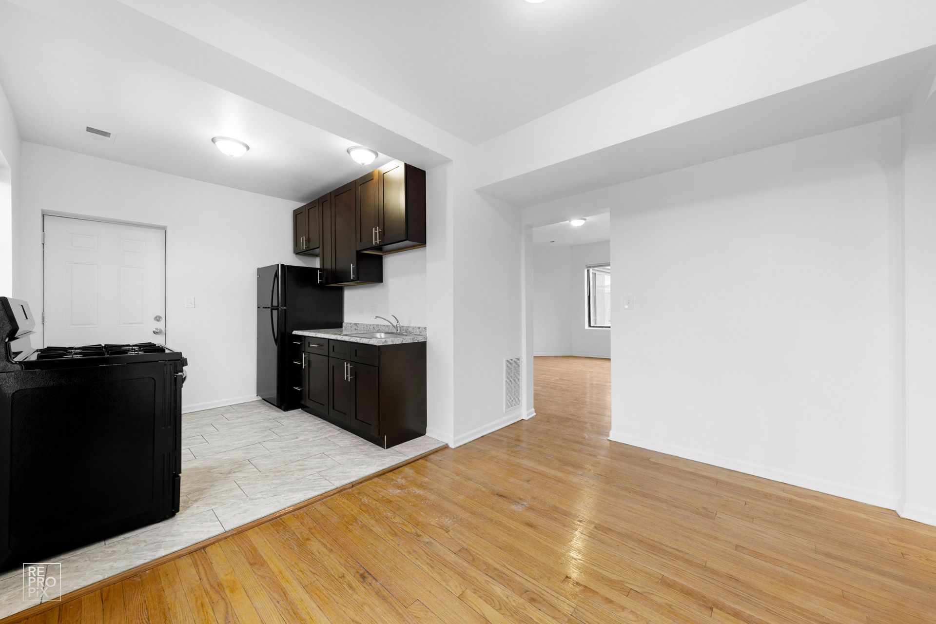 Kitchen with dark cabinets, black appliances, and wooden floors, connected to a living area with hardwood floors.