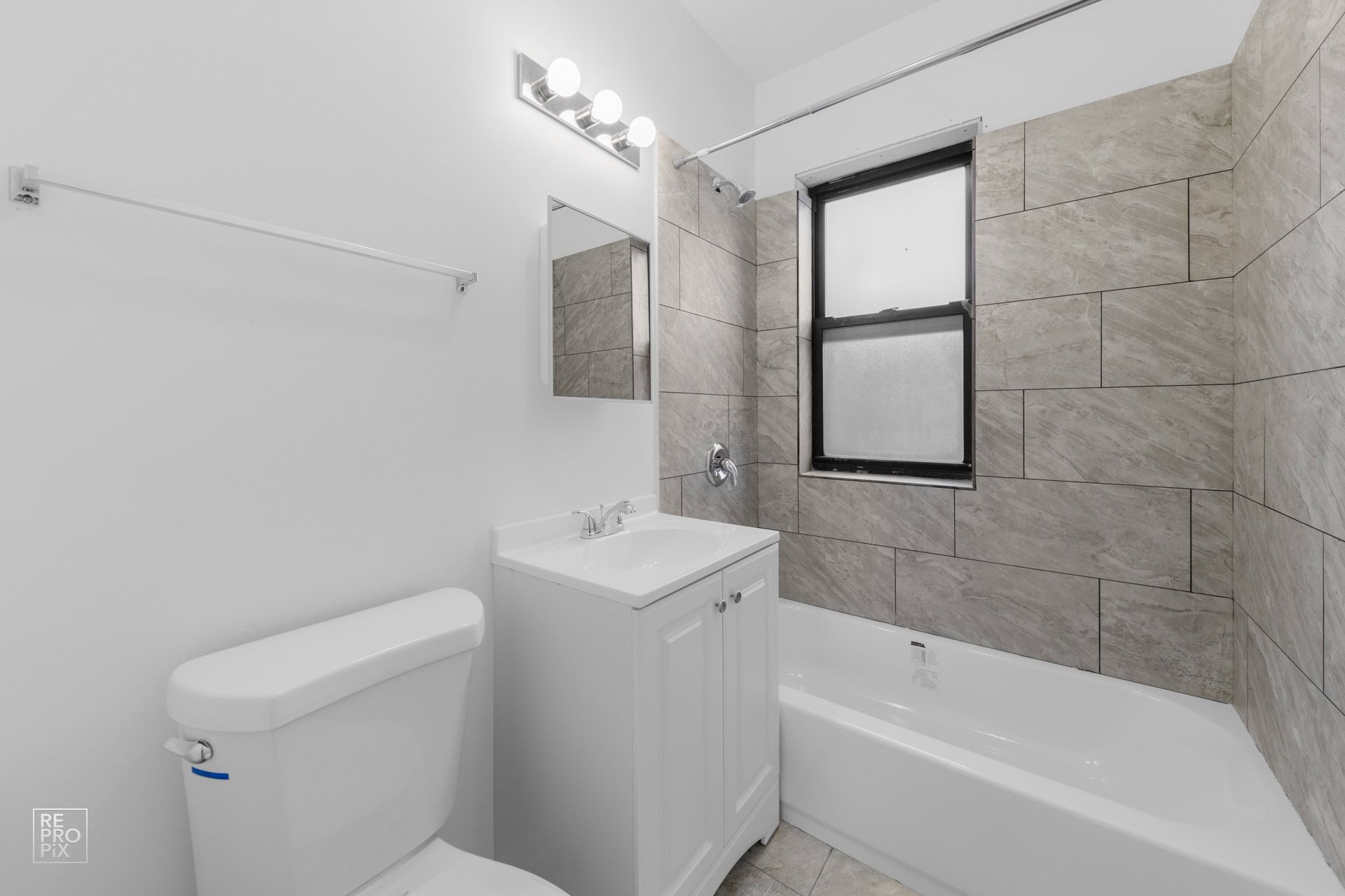 Small white bathroom with a toilet, sink, mirror, and bathtub. Light gray tiled wall.