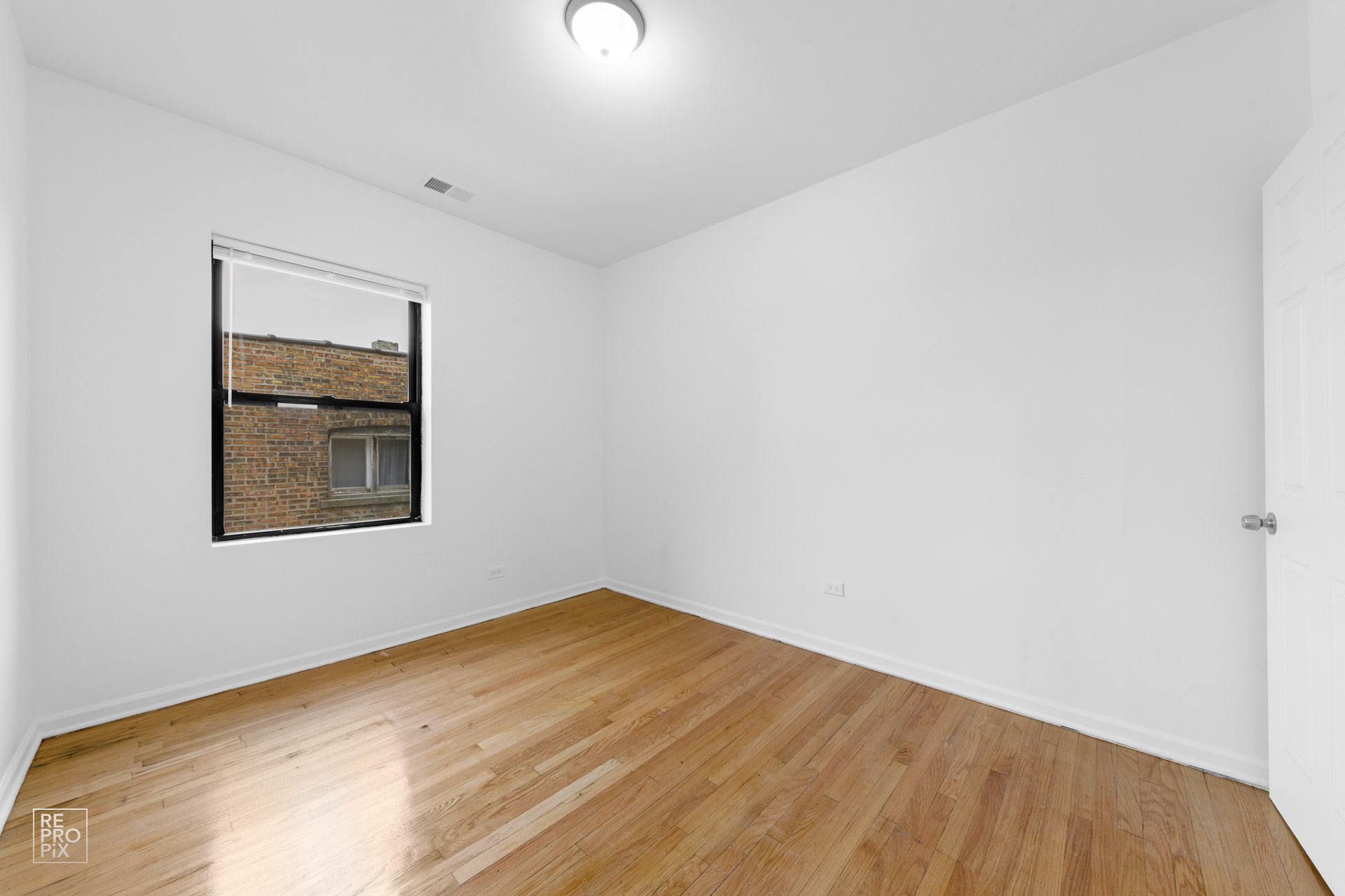 Empty room with wood floors, a window, and a closed door. White walls and ceiling with a single light fixture.