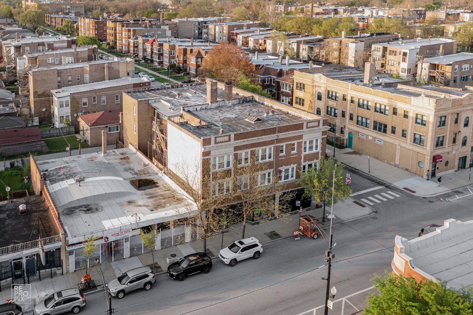 Aerial view of buildings on a city street; cars parked along the curb; trees with emerging leaves.