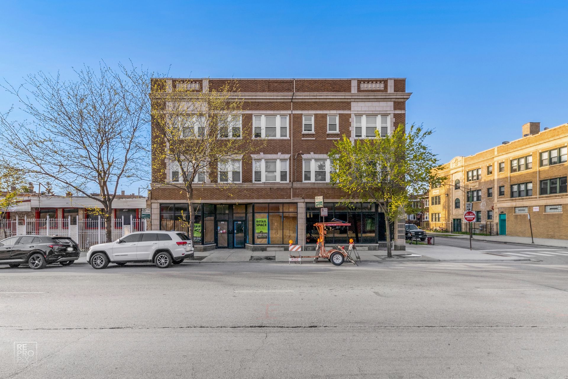 Brick building with storefronts on a city street. Cars are parked in front.