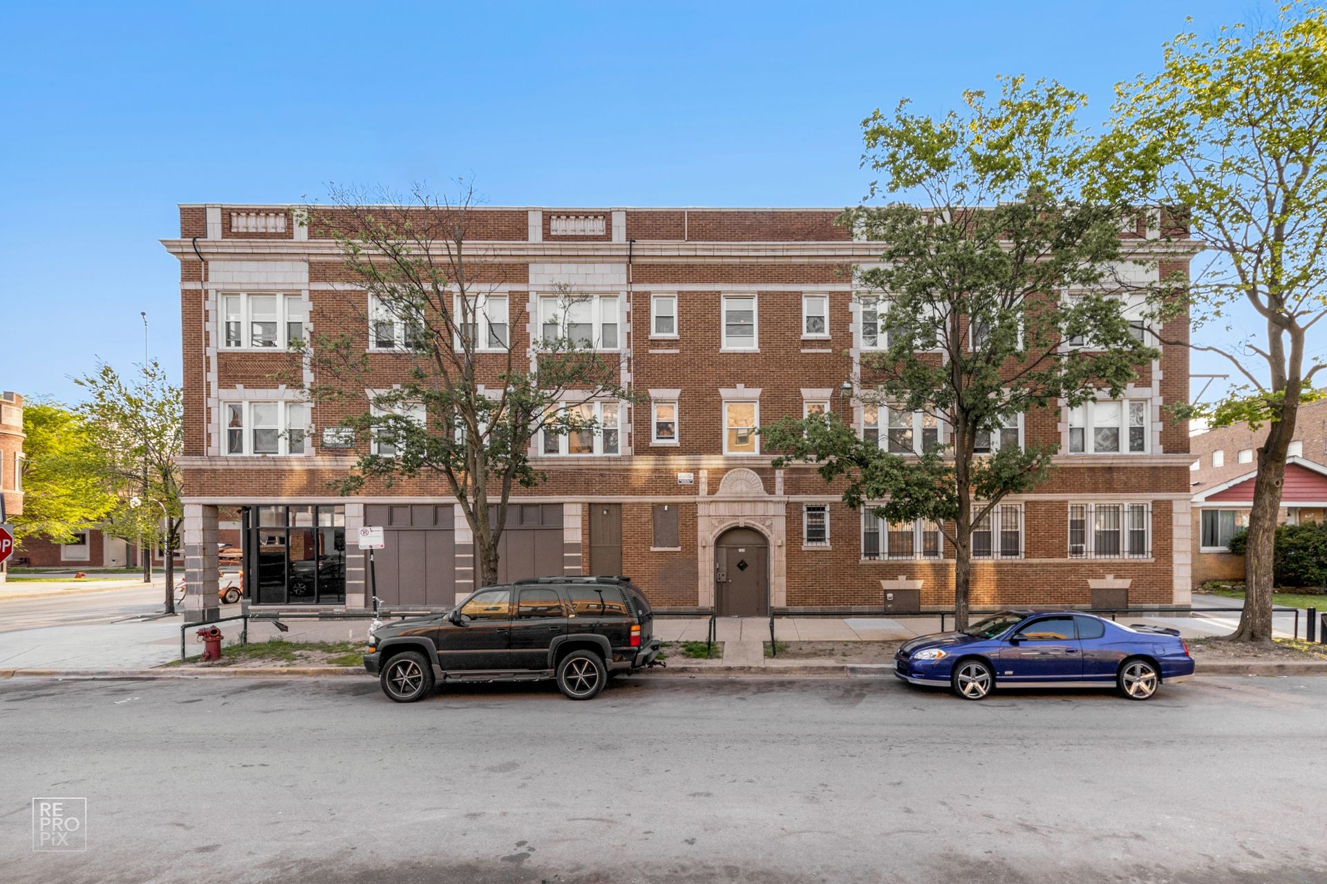 Brick apartment building with parked cars on the street. Brown, tan, and gray colors. Trees in front.