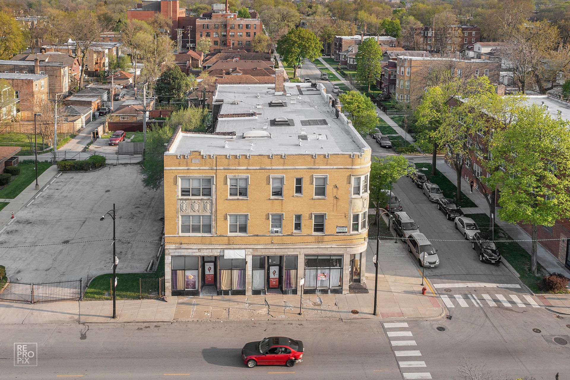Yellow building on a street corner, with empty lot and parked cars. A red car drives on the road.
