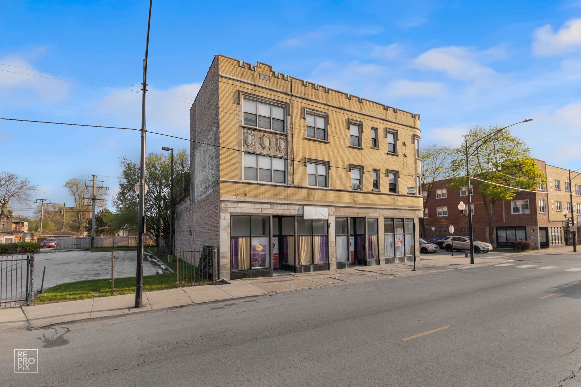 Three-story tan brick building with empty storefronts on a street. Cars parked on the right.