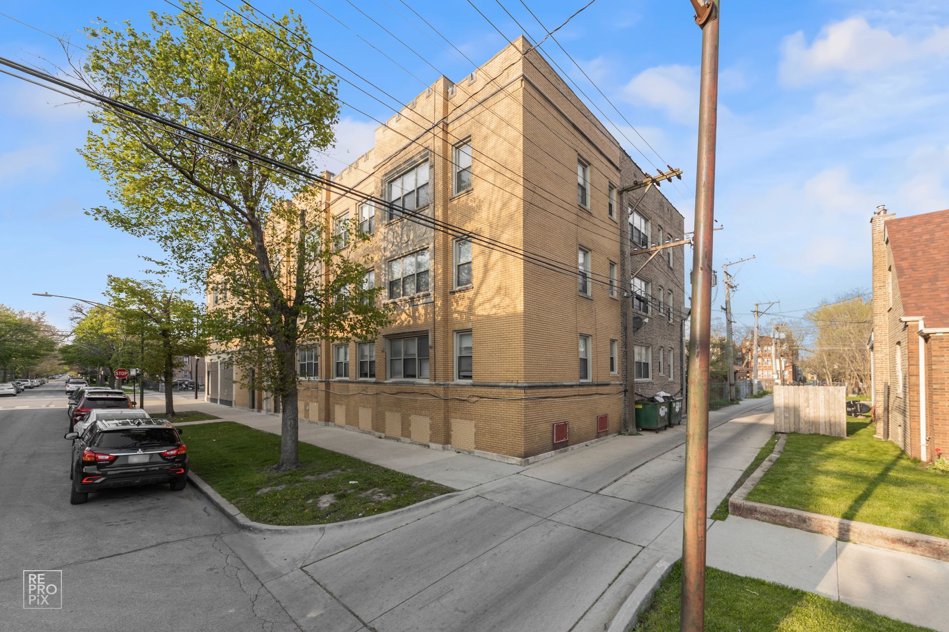 Corner view of a tan brick apartment building on a residential street with parked cars and a tree.