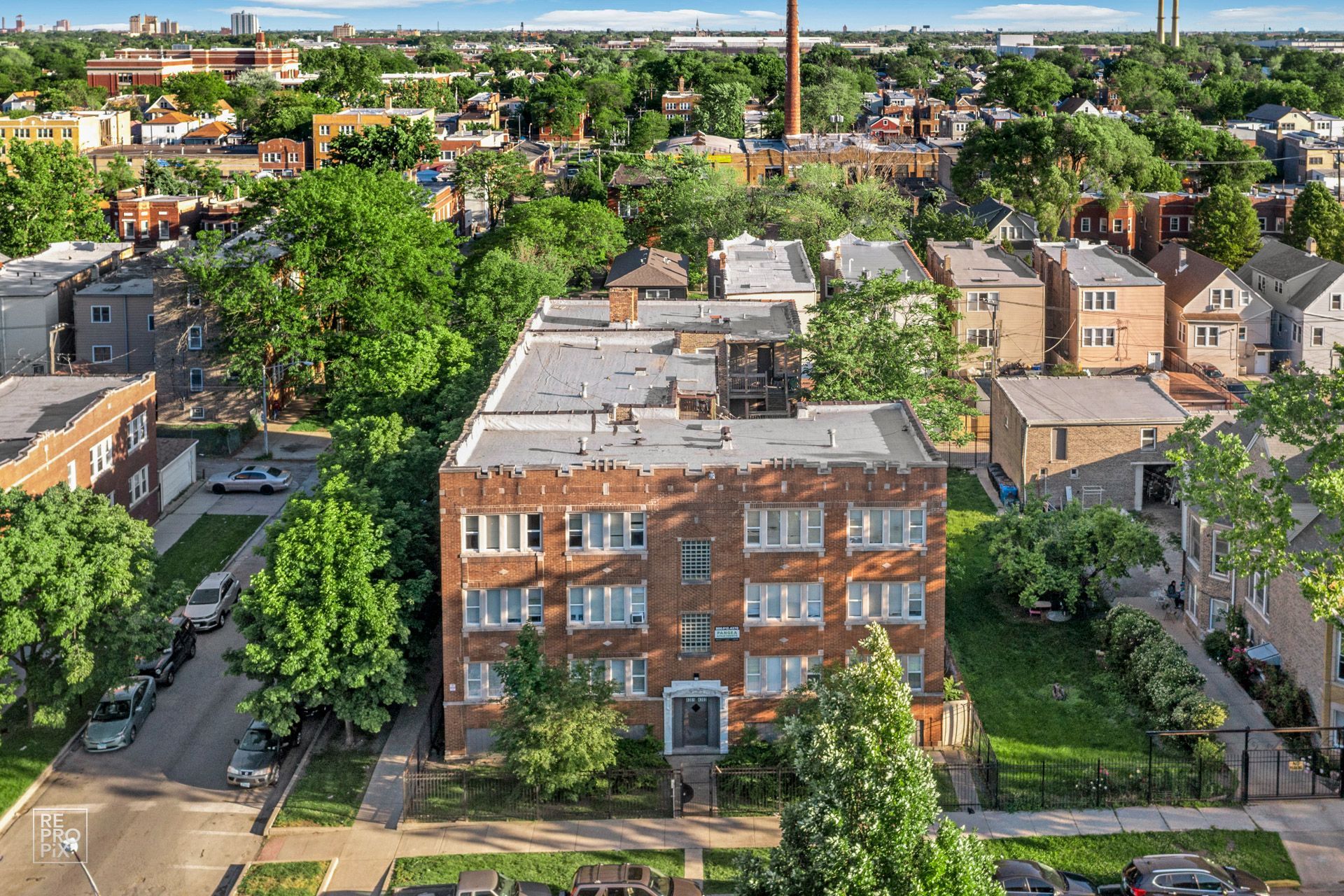 Aerial view of a three-story brick apartment building with a flat roof, surrounded by trees and residential buildings.