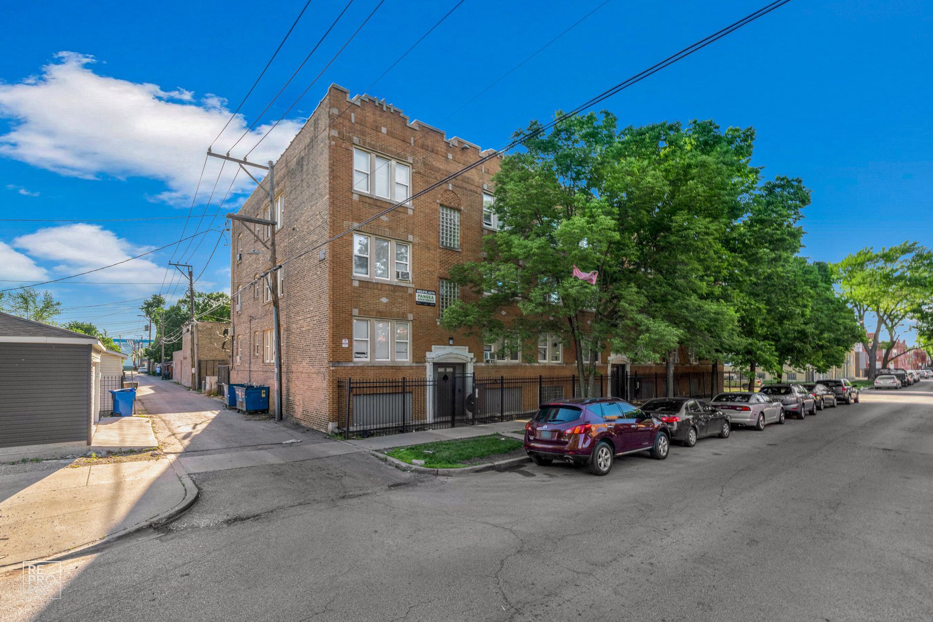 Brick apartment building on a street with parked cars and trees; blue sky in background.