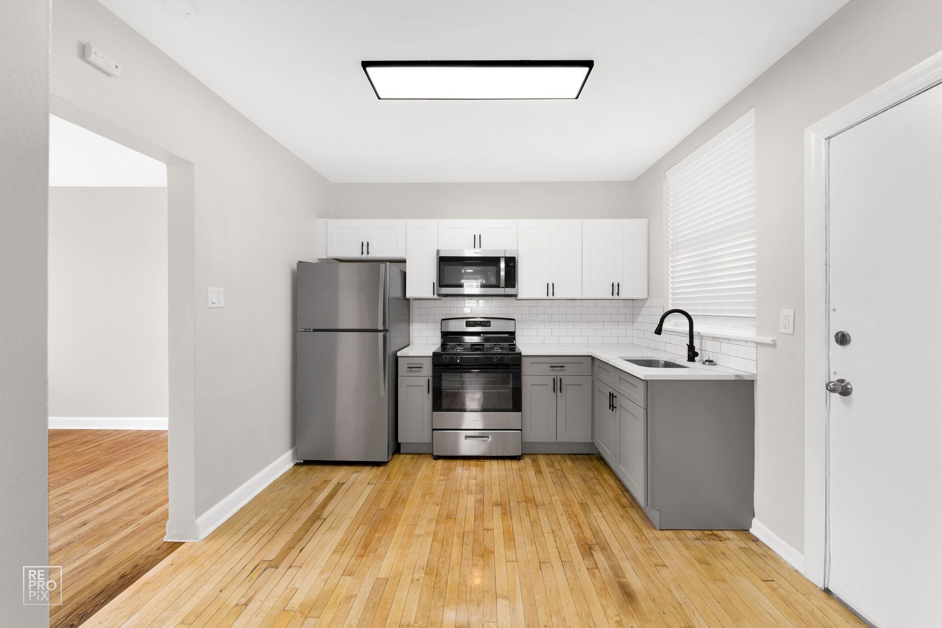 Kitchen with stainless steel appliances, white and gray cabinets, light wood floor, and doorway to another room.