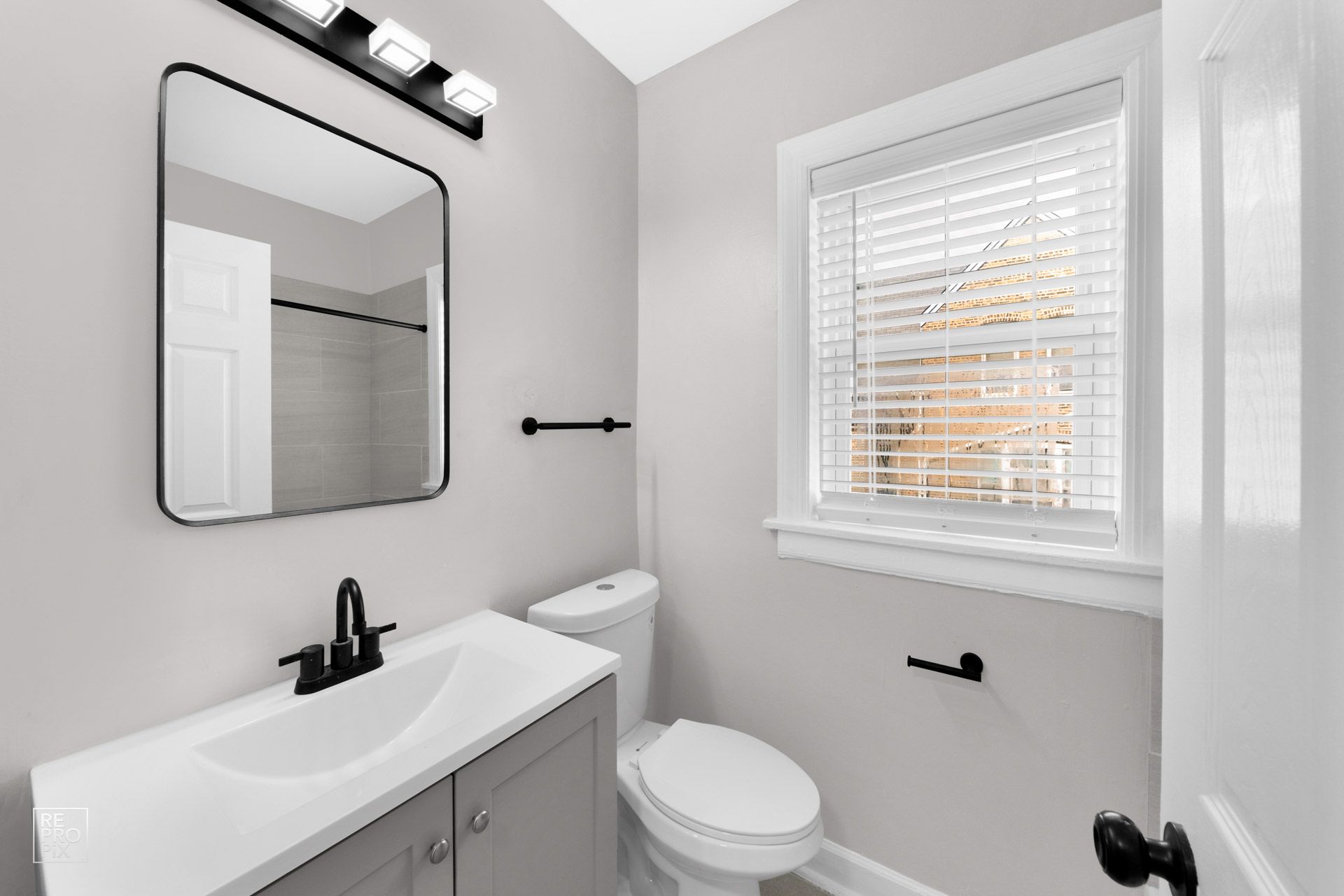 Gray bathroom with vanity, toilet, and window with blinds. Black fixtures and trim.