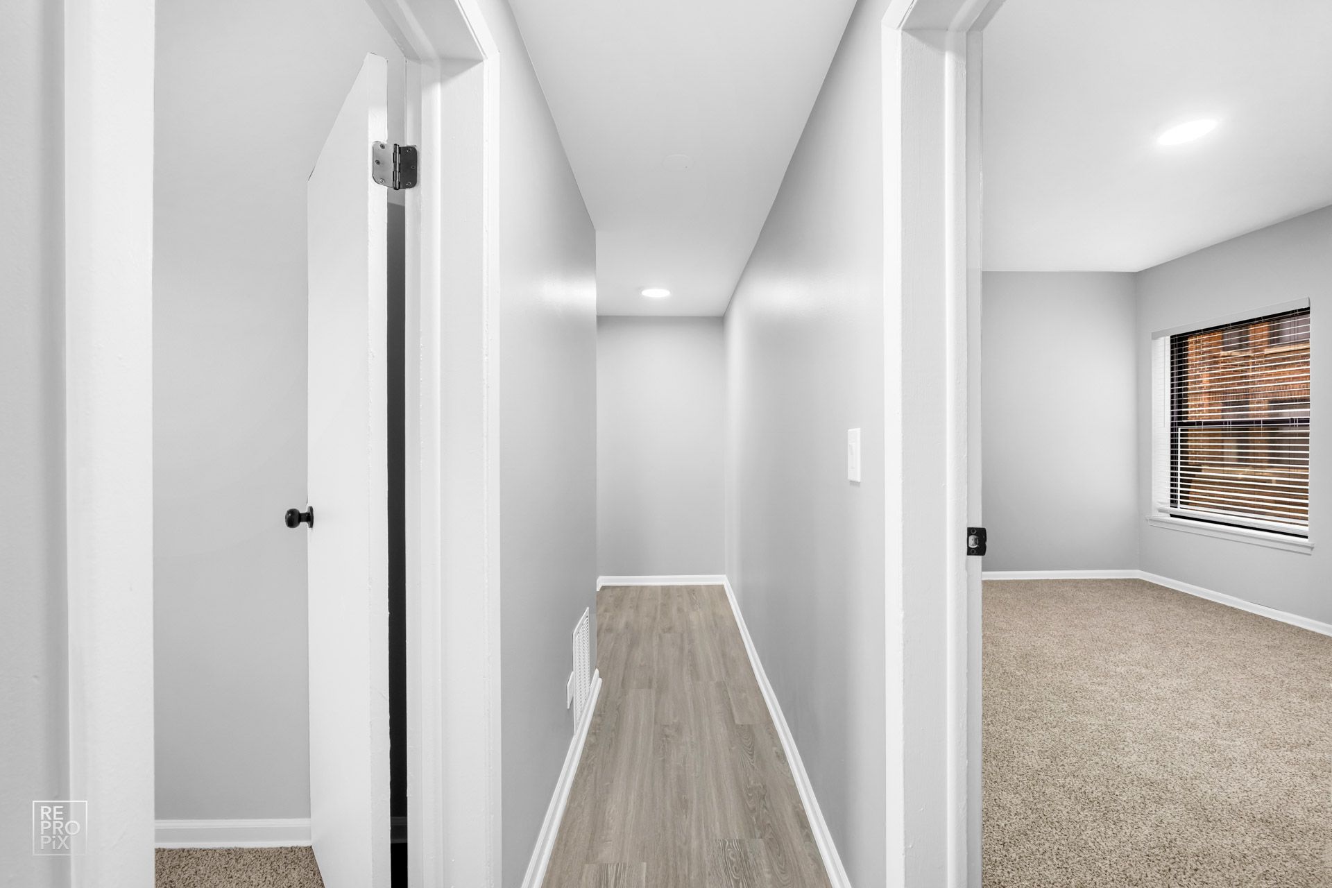 Narrow hallway with gray walls, light wood-look flooring, and doors on either side, one leading to a room with carpet and a window.