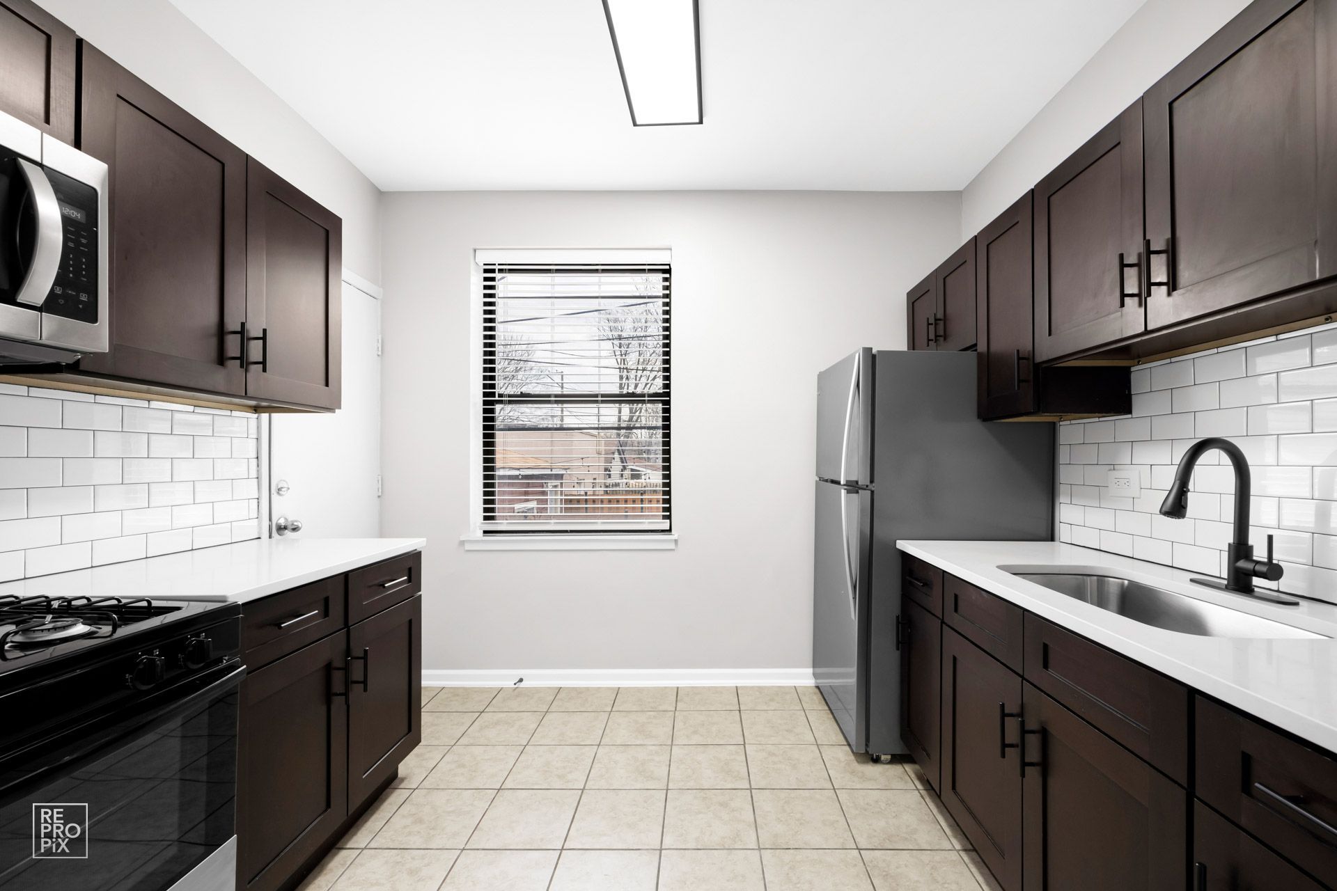 Modern kitchen with dark brown cabinets, white countertops, stainless steel appliances, and a window.
