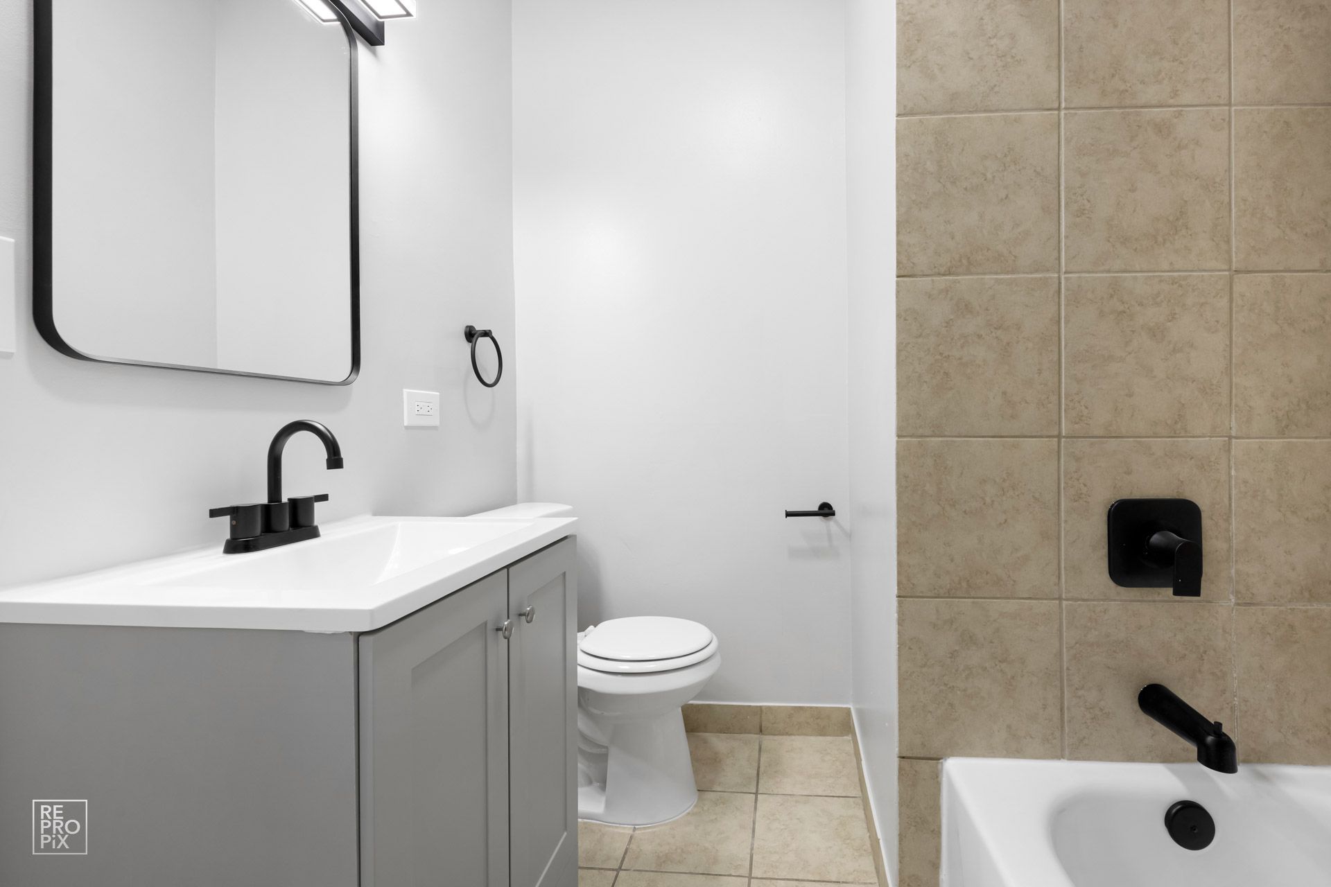 Bathroom with gray vanity, black fixtures, white toilet, and beige tiled shower.