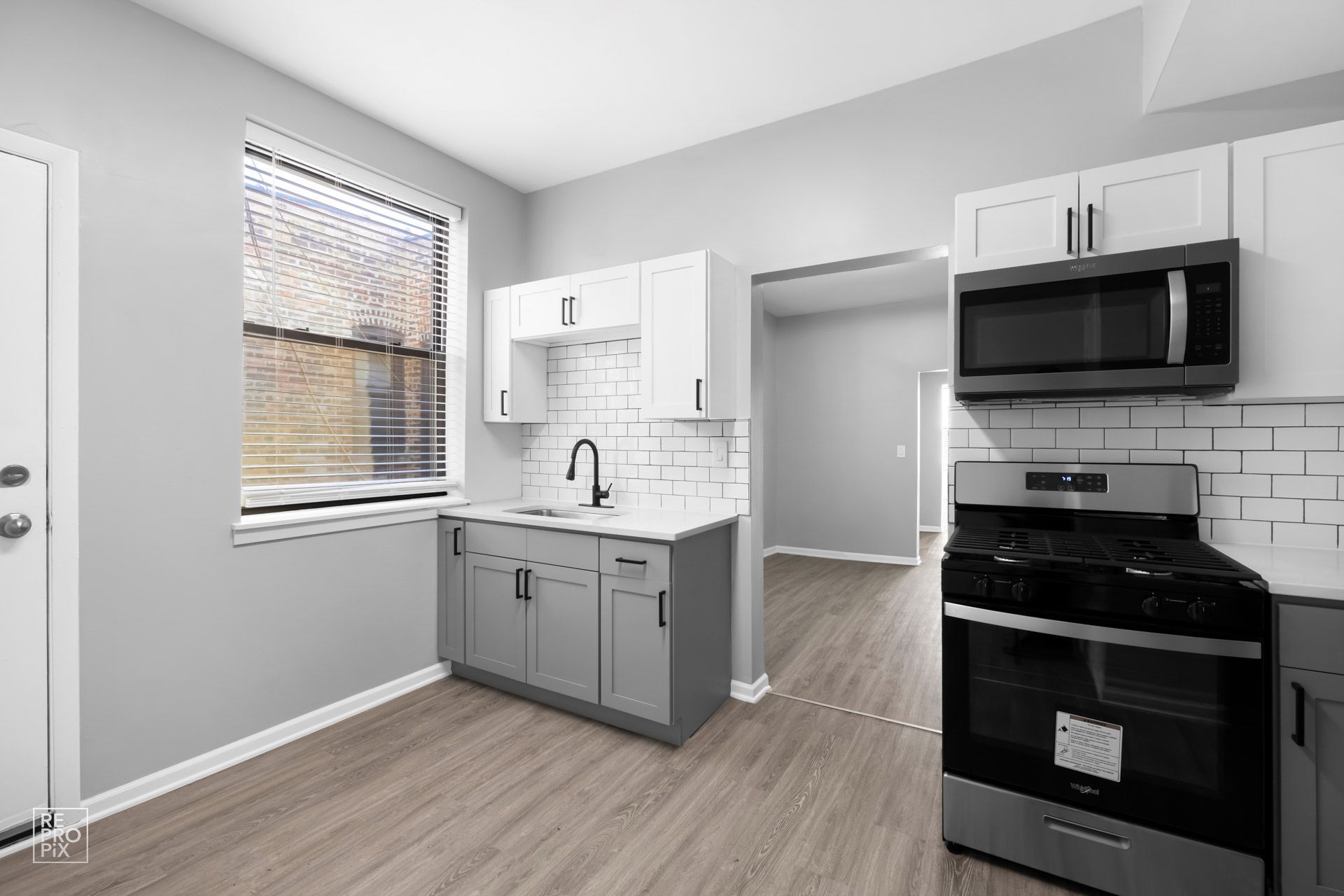 Kitchen with gray and white cabinets, black appliances, window with blinds, and wood-look flooring.