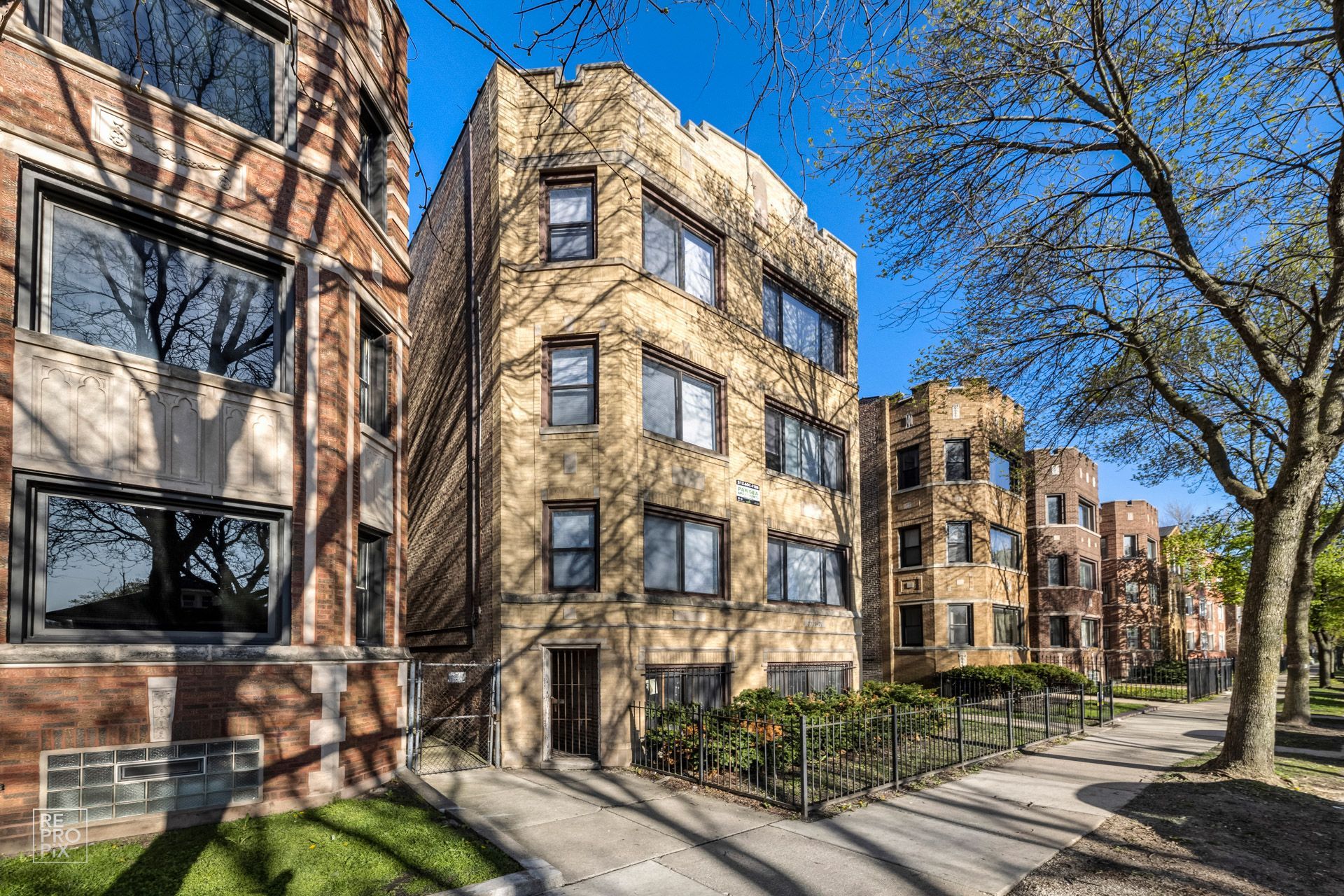 Multi-story tan brick apartment building with several windows and a small front yard, under a blue sky.