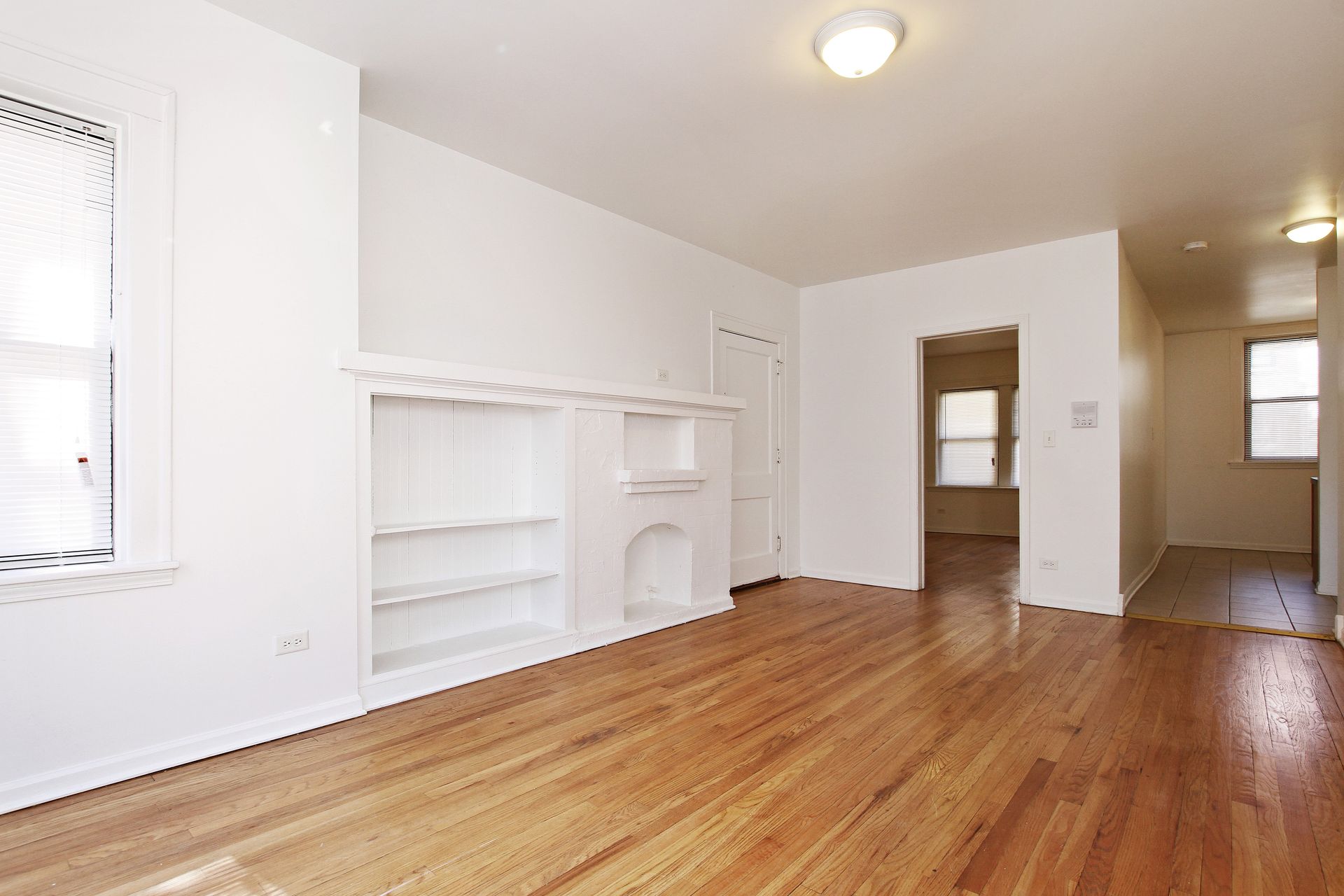 Empty, bright living room with hardwood floors, white walls, built-in shelving, and doorways.