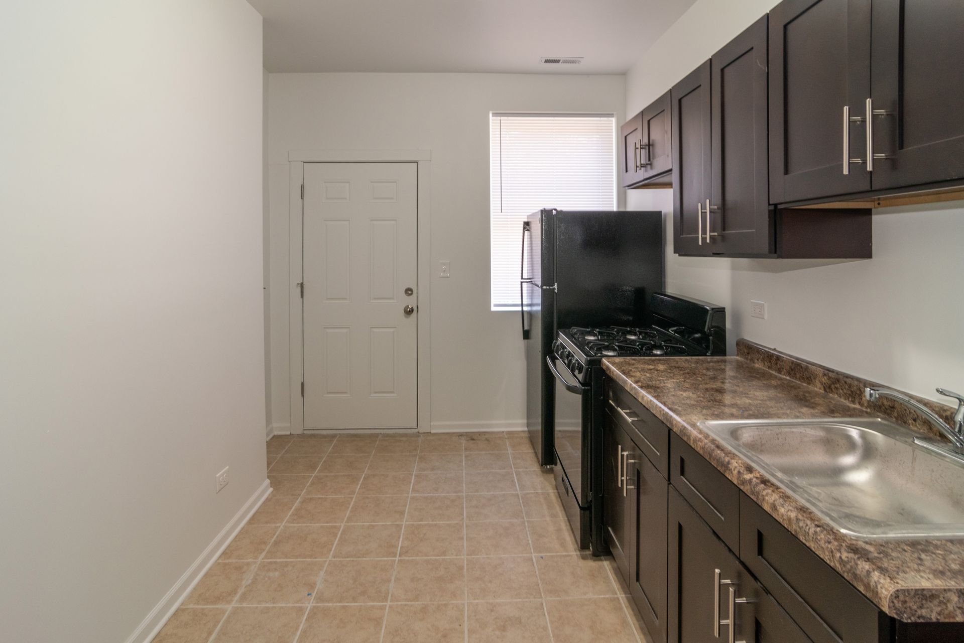 Kitchen with dark cabinets, black appliances, a door, and a window with blinds.