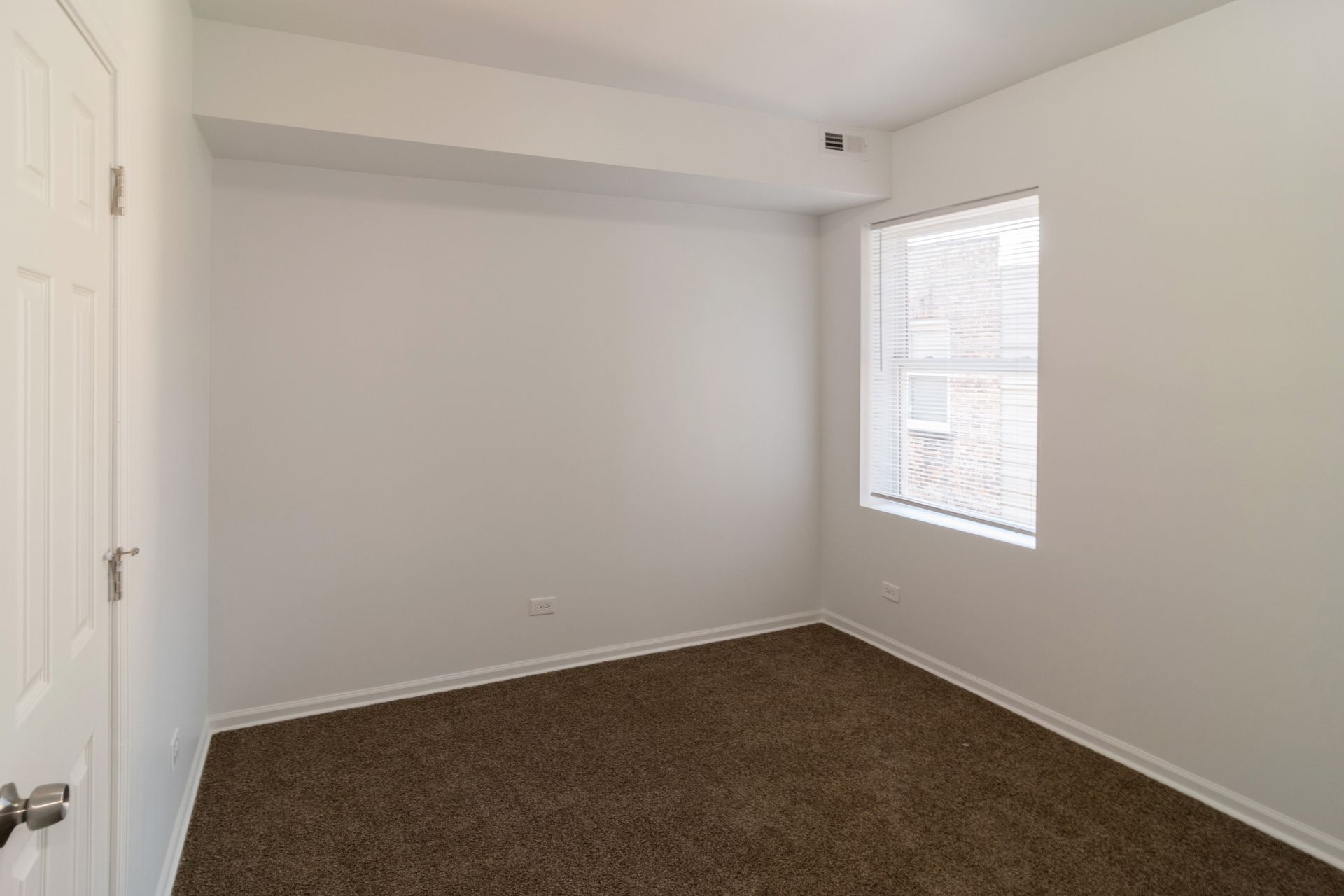 Empty room with white walls, brown carpet, window, and closed door.
